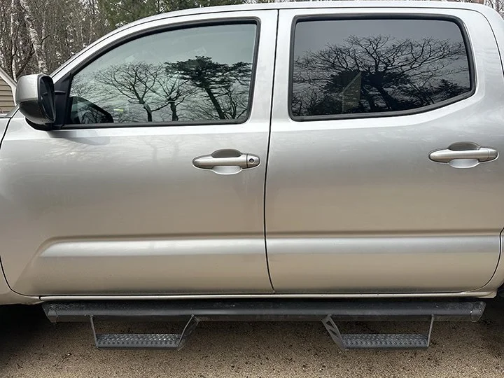 Silver pickup truck with sliding rear doors and running boards, parked outdoors with leafless trees and cloudy sky reflected in the windows.