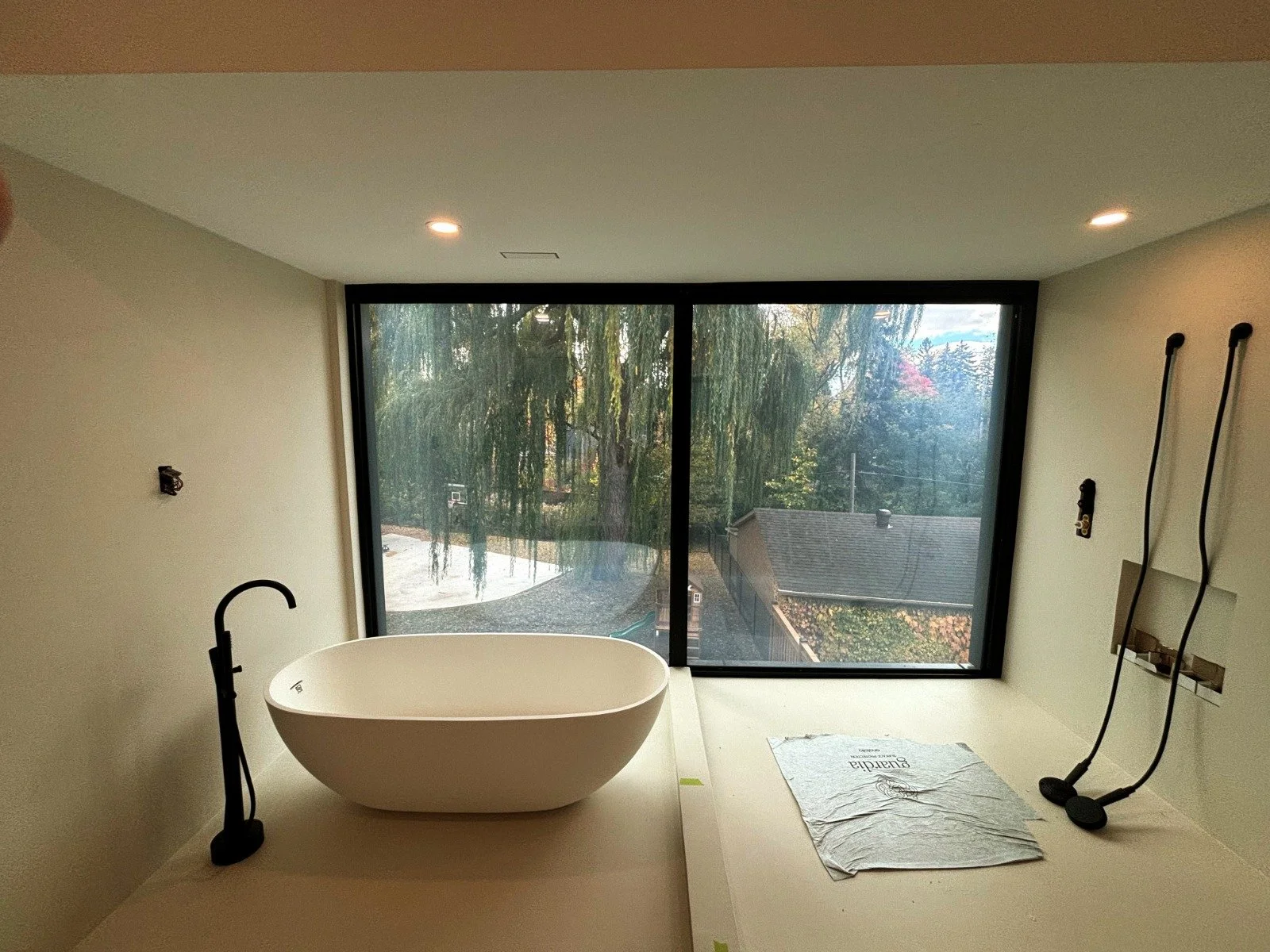 Modern bathroom with a standalone bathtub in front of large window showing trees and a house roof outside, with tools and tape on the surface, and recessed ceiling lights.