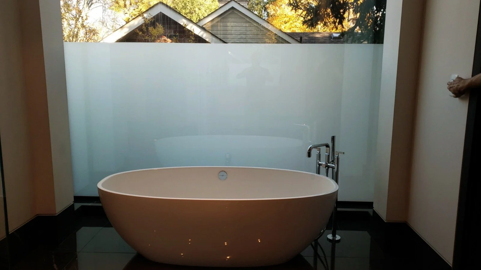 A modern bathroom features a white oval soaking tub with a chrome faucet and a frosted glass window or wall behind it, with an outdoor view of trees and the roof of neighboring houses.