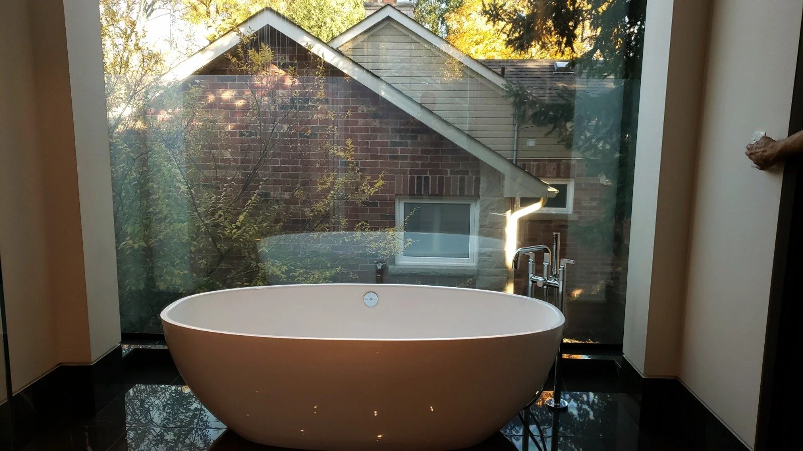 A modern white freestanding bathtub placed in front of a large window, with a view of a brick house and trees outside.