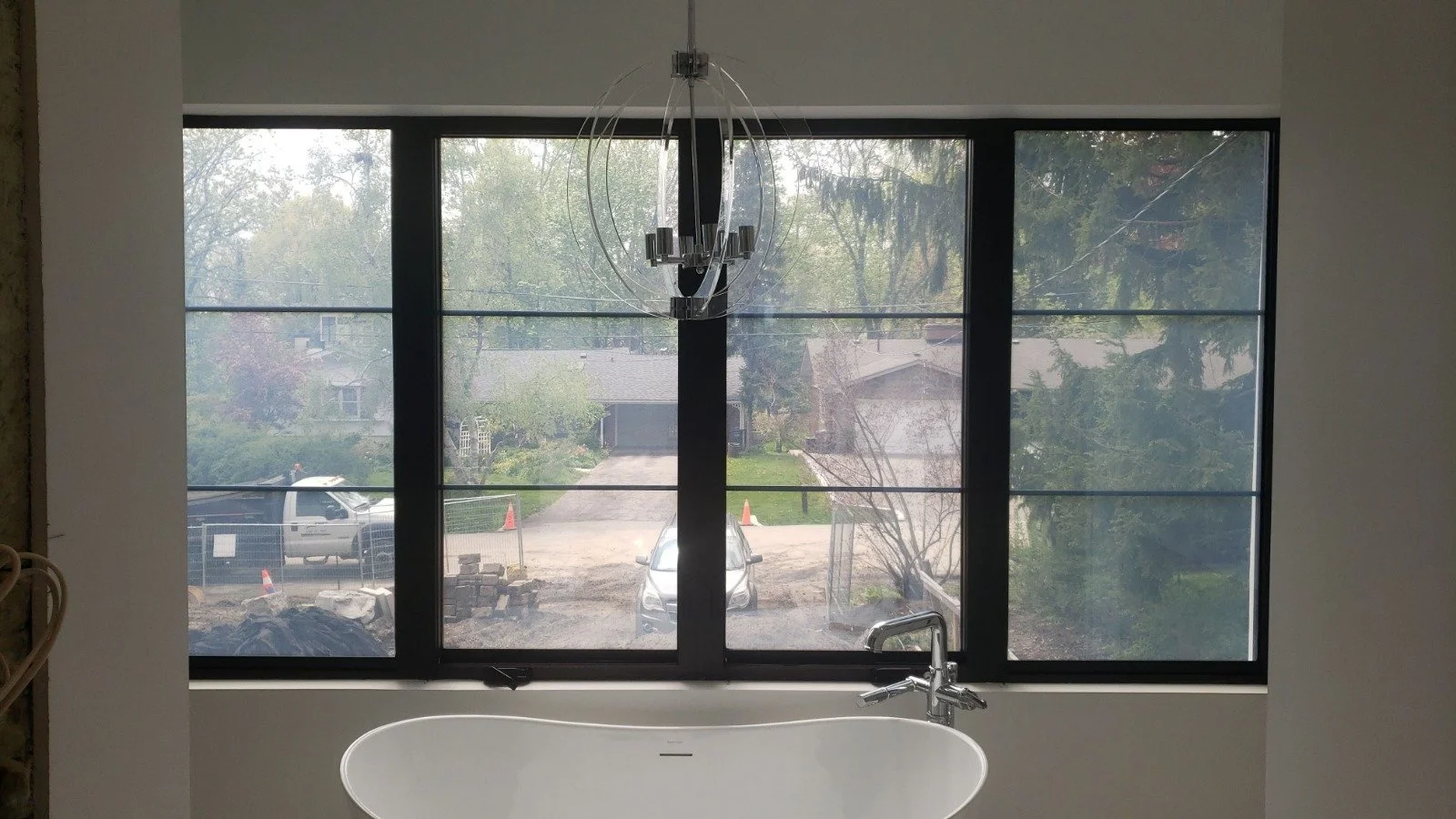 Bathroom with large window overlooking a driveway and trees, bathtub below the window, modern chandelier hanging from the ceiling.