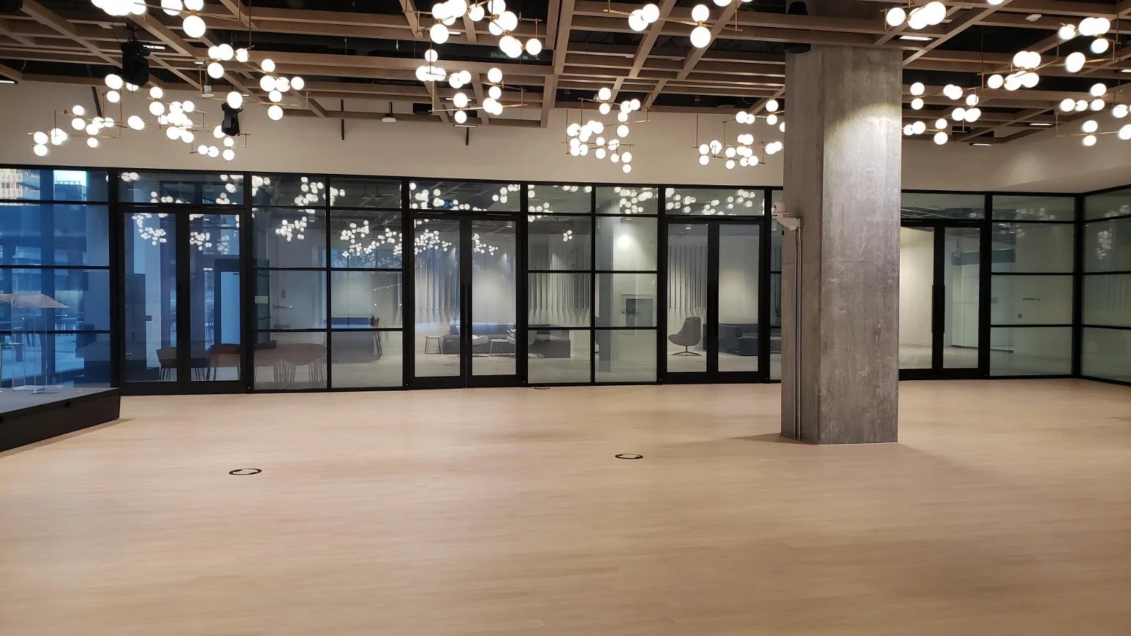 Empty modern office space with large glass walls, wood flooring, and decorative ceiling lights.