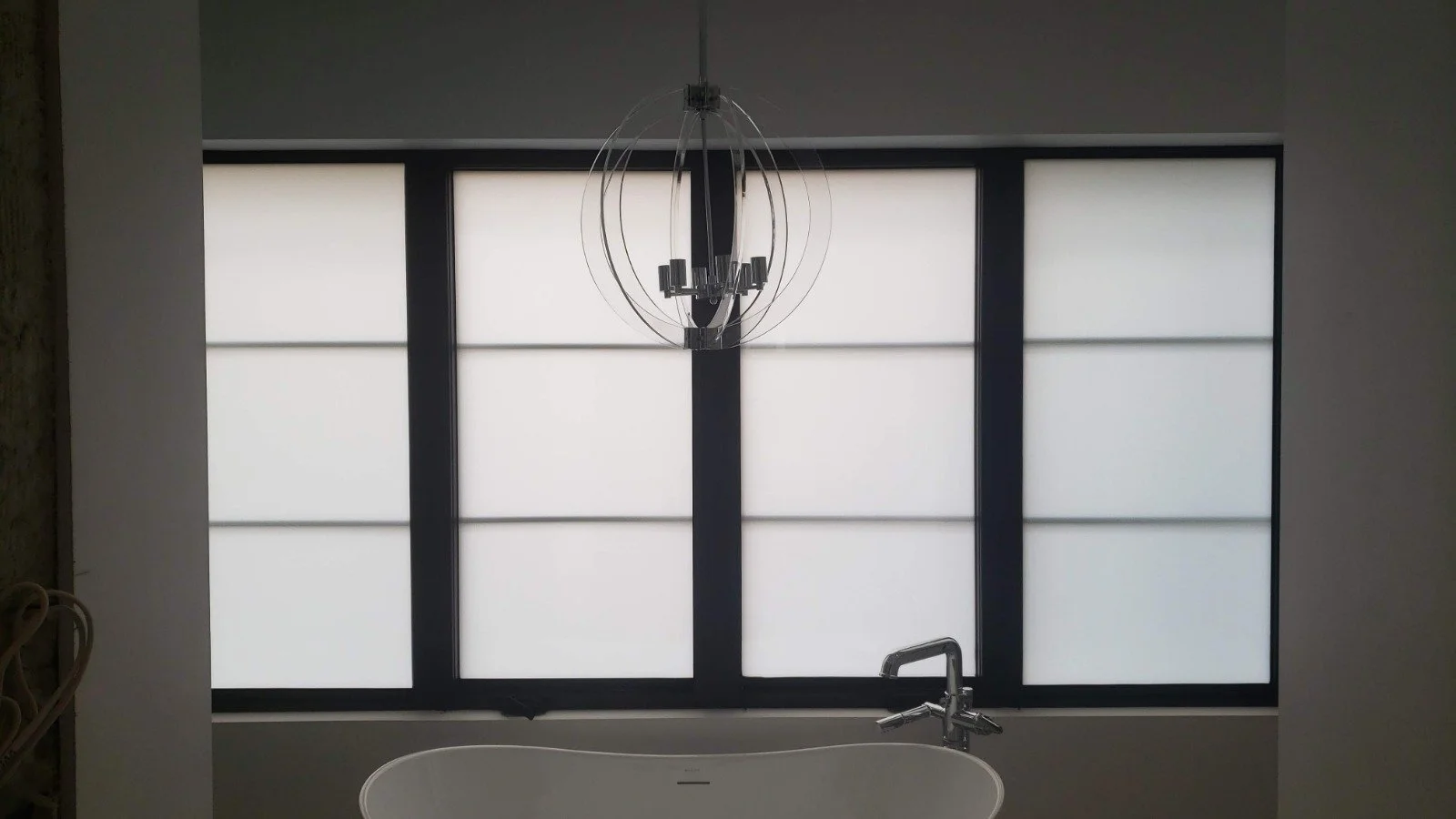 Modern bathroom with a black-framed window covered by frosted glass, a contemporary chandelier hanging from the ceiling, a white bathtub with a silver faucet in the foreground.