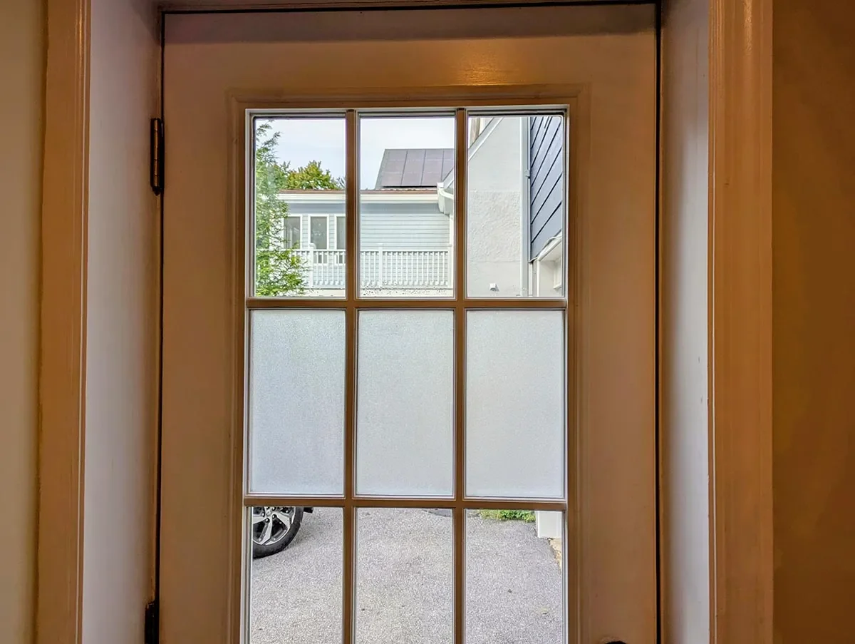 View through a door with a window showing a neighborhood scene with a car, houses, and trees outside.