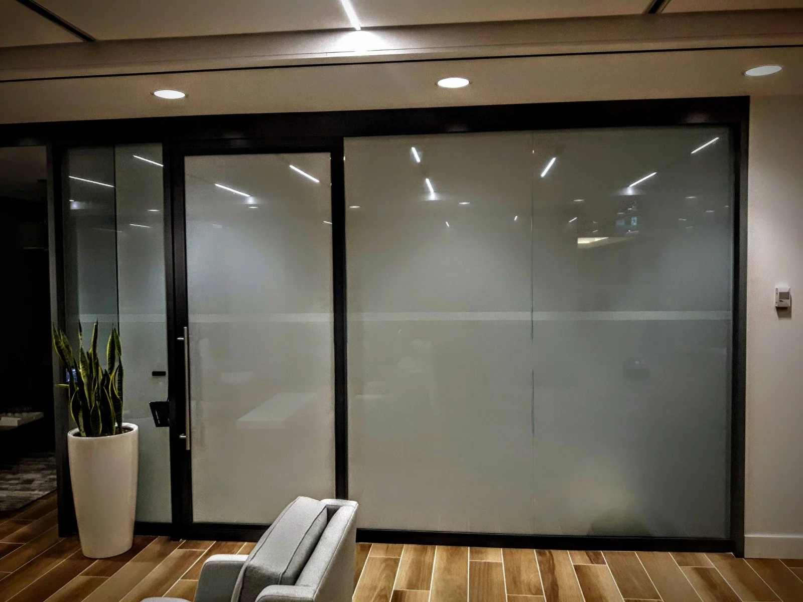 Frosted glass doors with black metal frame in an office setting, with a potted plant, gray chair, wooden floor, and ceiling lights reflected on the glass.
