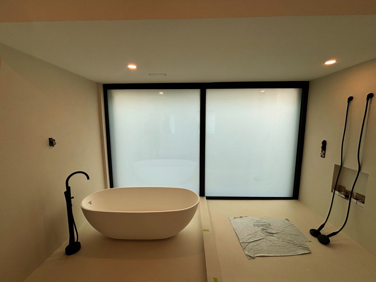 A bathroom under construction with a white bathtub, a black faucet, two wall-mounted black showerheads, and frosted glass windows.
