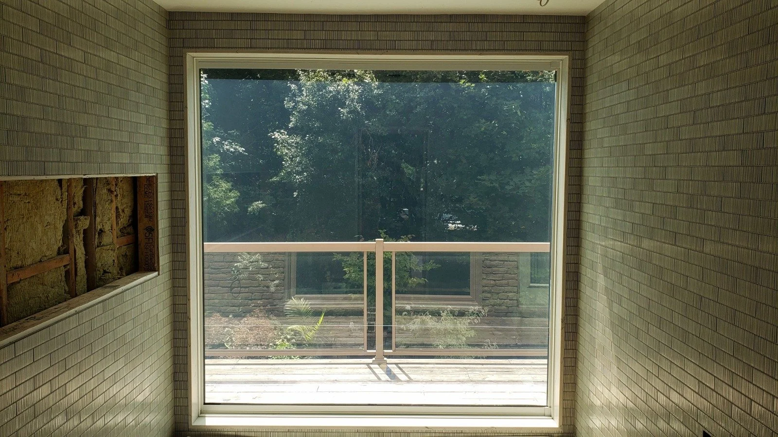 Interior view of a room with beige brick walls, a large window with a view of a green outdoor space, and a balcony with a railing.