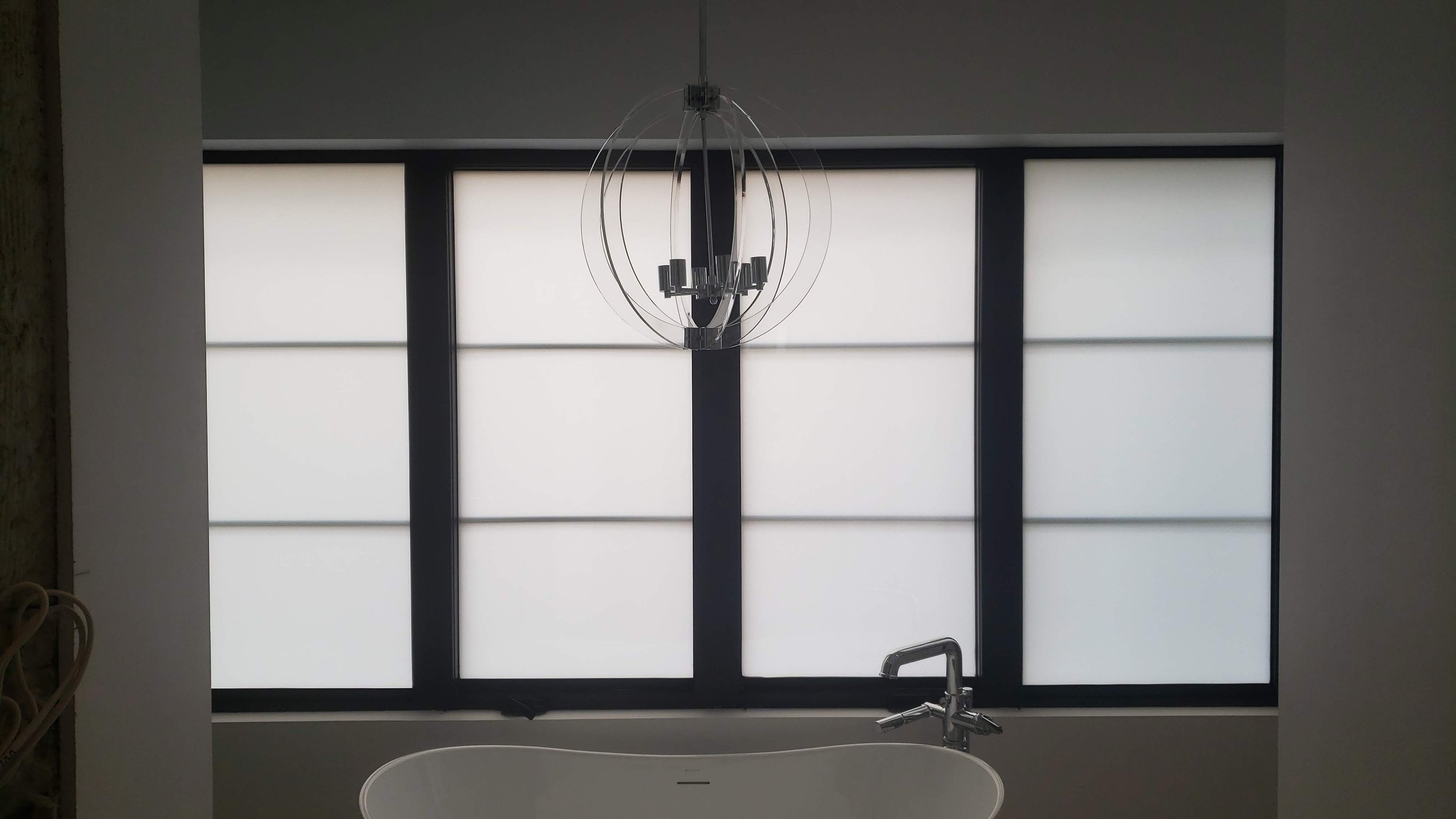 Modern bathroom with a large window with frosted glass, a white bathtub in front of the window, and a contemporary chandelier hanging from the ceiling.