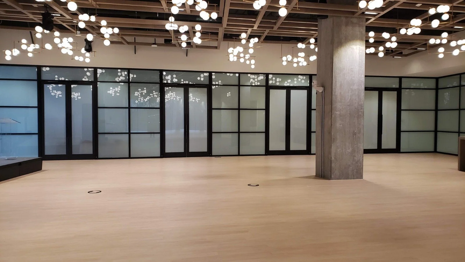 Empty modern interior space with wooden flooring, glass walls with black frames, a large concrete column, and ceiling lights resembling small glowing orbs.