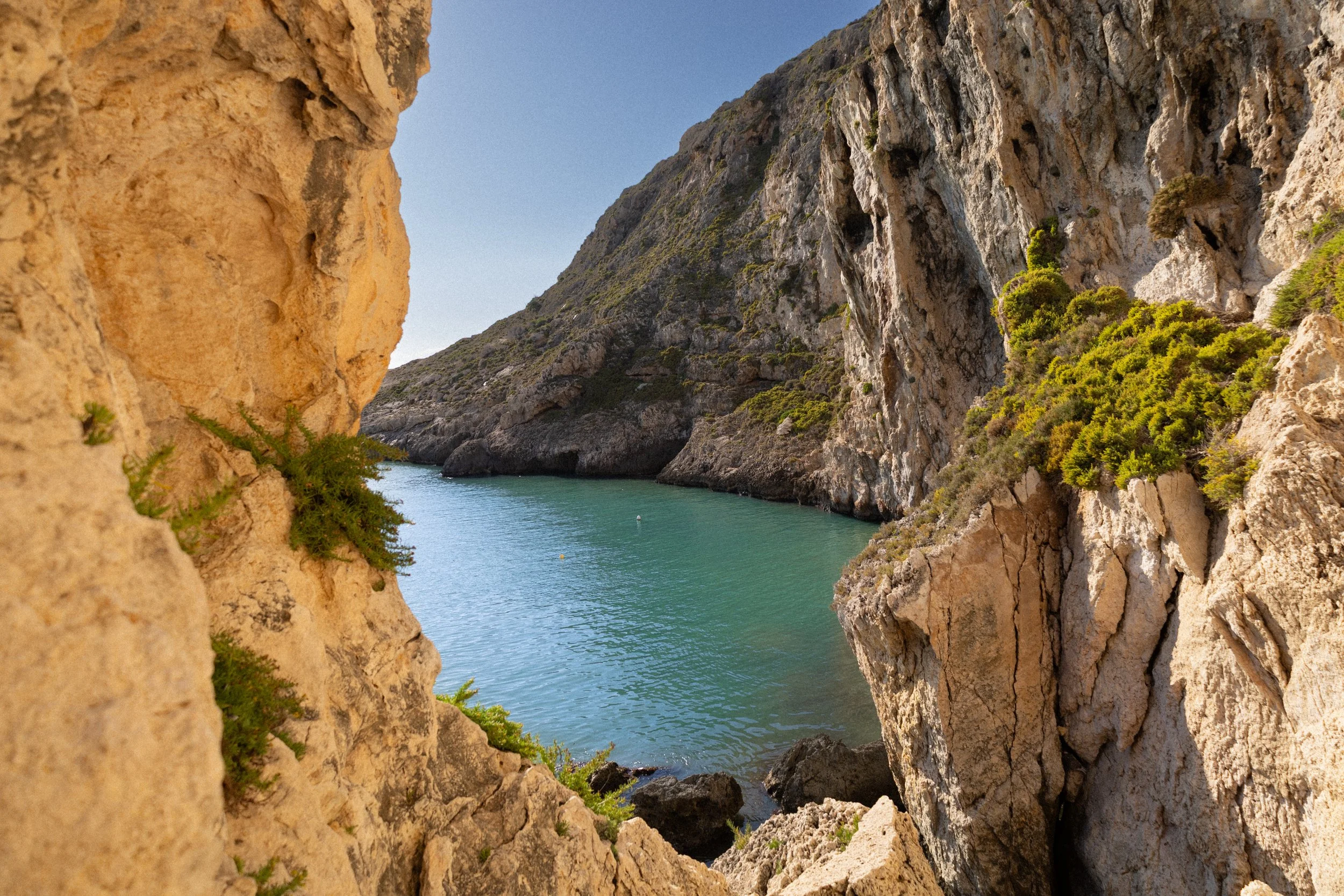 View through rocky cliffs of a turquoise water inlet in Malta, with steep, rocky hills in the background under a clear blue sky.