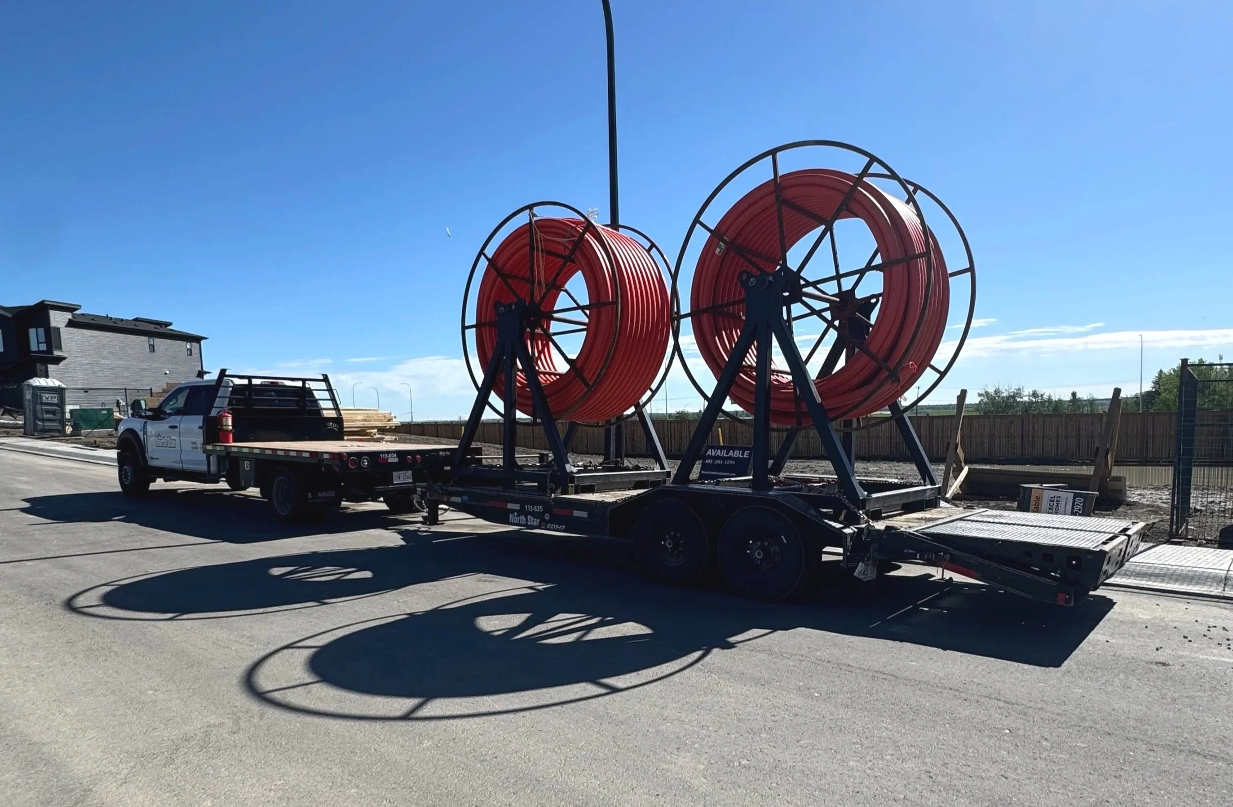 Flatbed truck carrying large orange fire hoses with black supports, parked on a street under a clear blue sky.