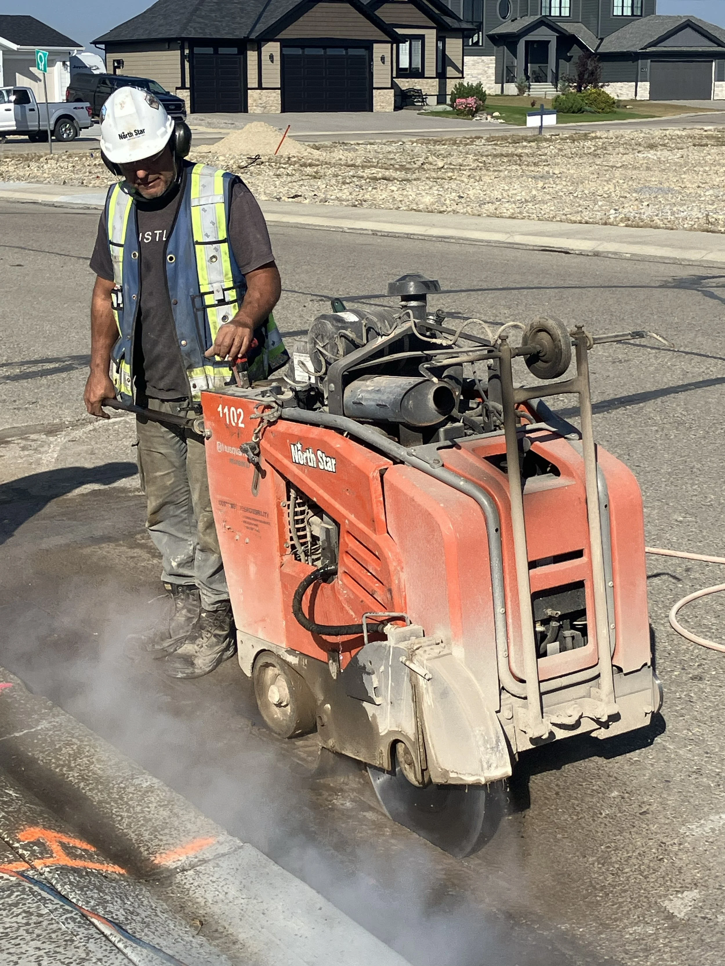 Construction worker operating a small asphalt paving machine on a residential street during daytime. The worker is wearing a white helmet and a safety vest, with houses and parked cars visible in the background.