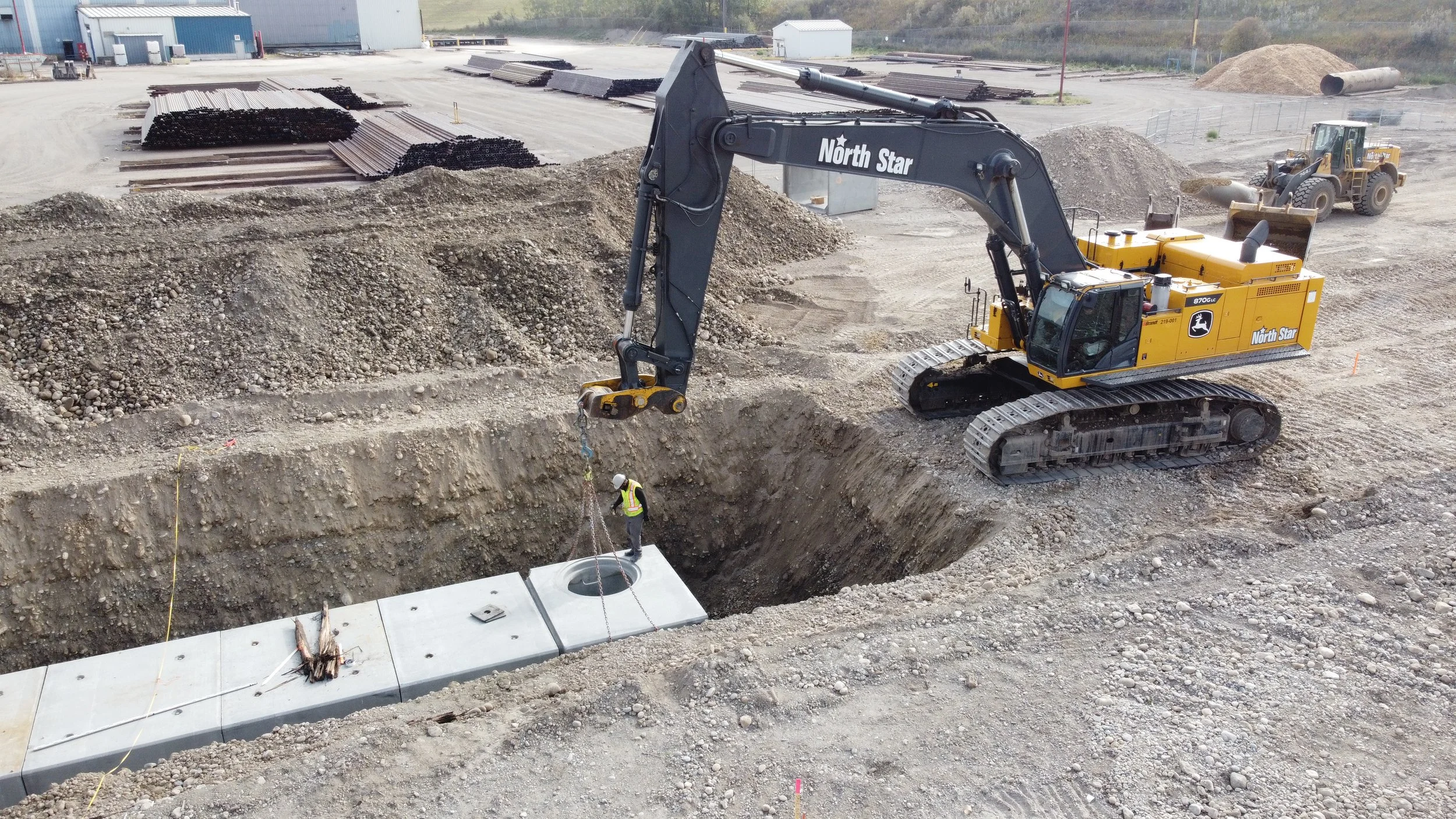 A construction site with a large excavator from North Star company lifting pre-cast concrete panels, with a worker nearby guiding the panels into place. There are piles of dirt and construction materials, with industrial buildings in the background.