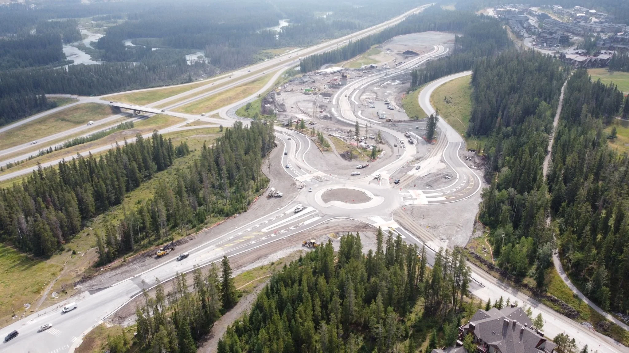 Aerial view of a highway construction site surrounded by dense trees, with roads, vehicles, and partially built roadways.