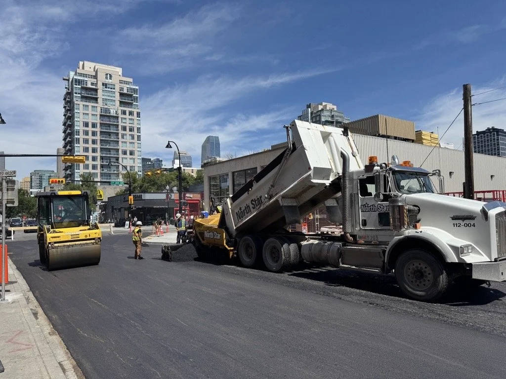 Construction workers paving a street in an urban area with high-rise buildings in the background.