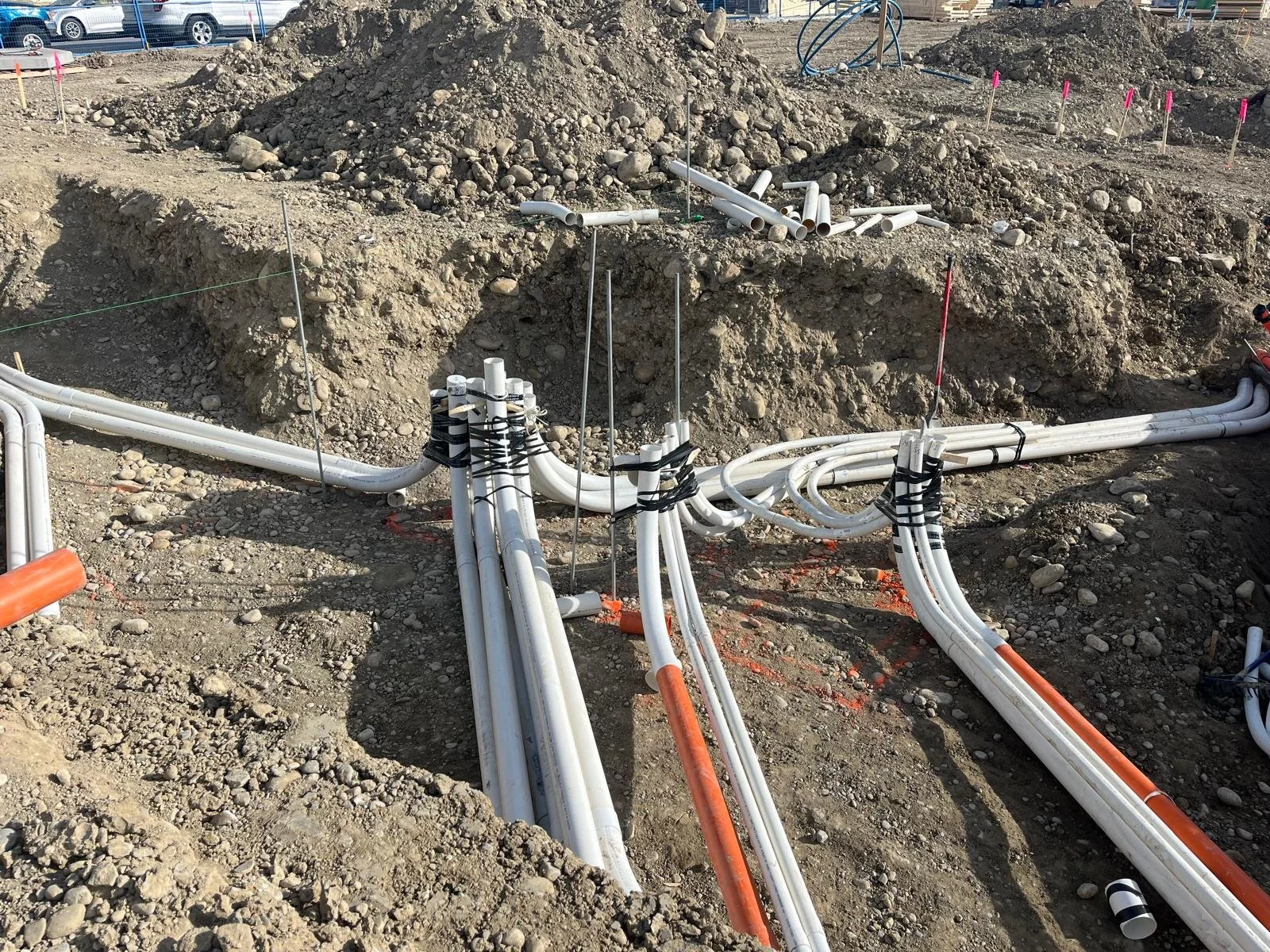 Construction site with multiple white and orange PVC pipes laid out on the ground, some connected and some loose, with mounds of dirt and gravel in the background.