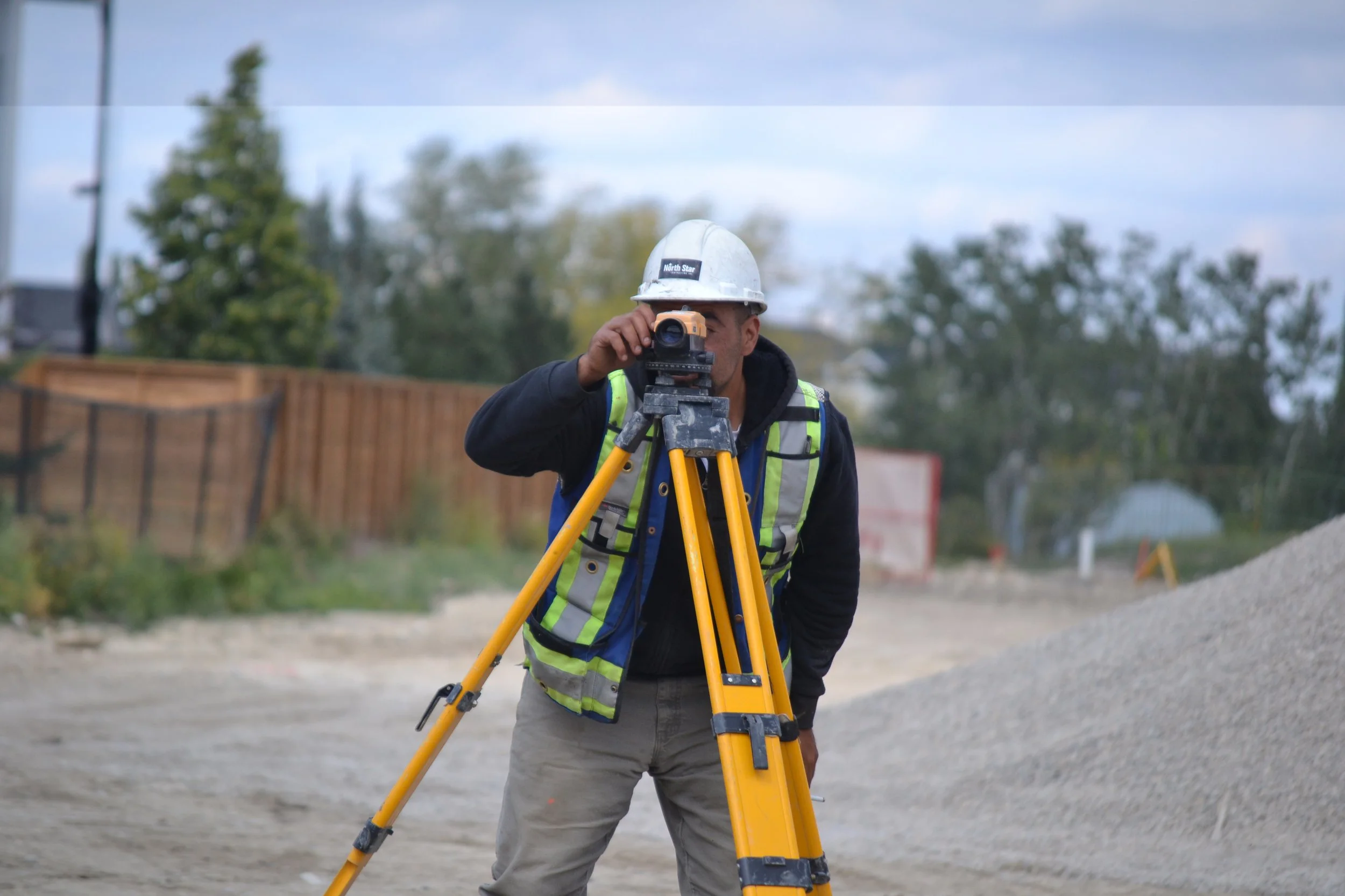 A construction worker wearing a white safety helmet and a reflective safety vest operating surveying equipment at a construction site.