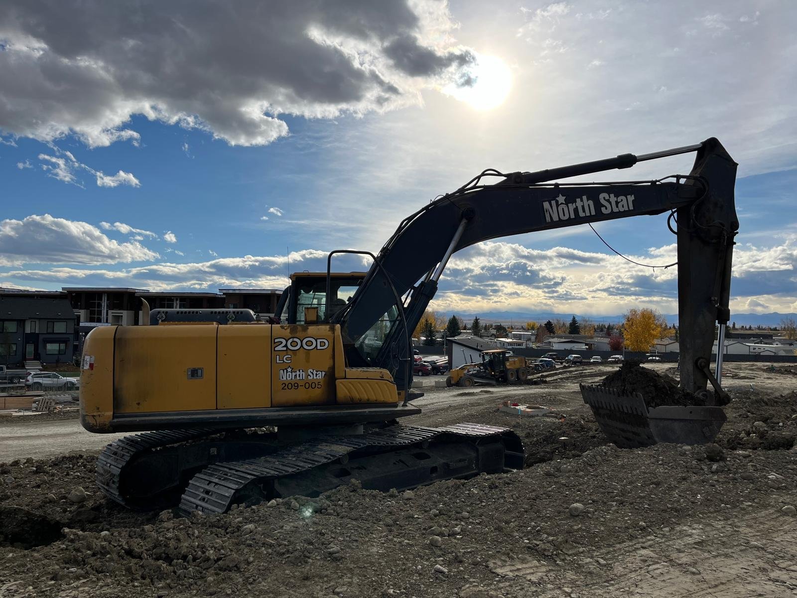 A yellow and black excavator on a construction site with buildings and trees in the background, under a partly cloudy sky with the sun shining.