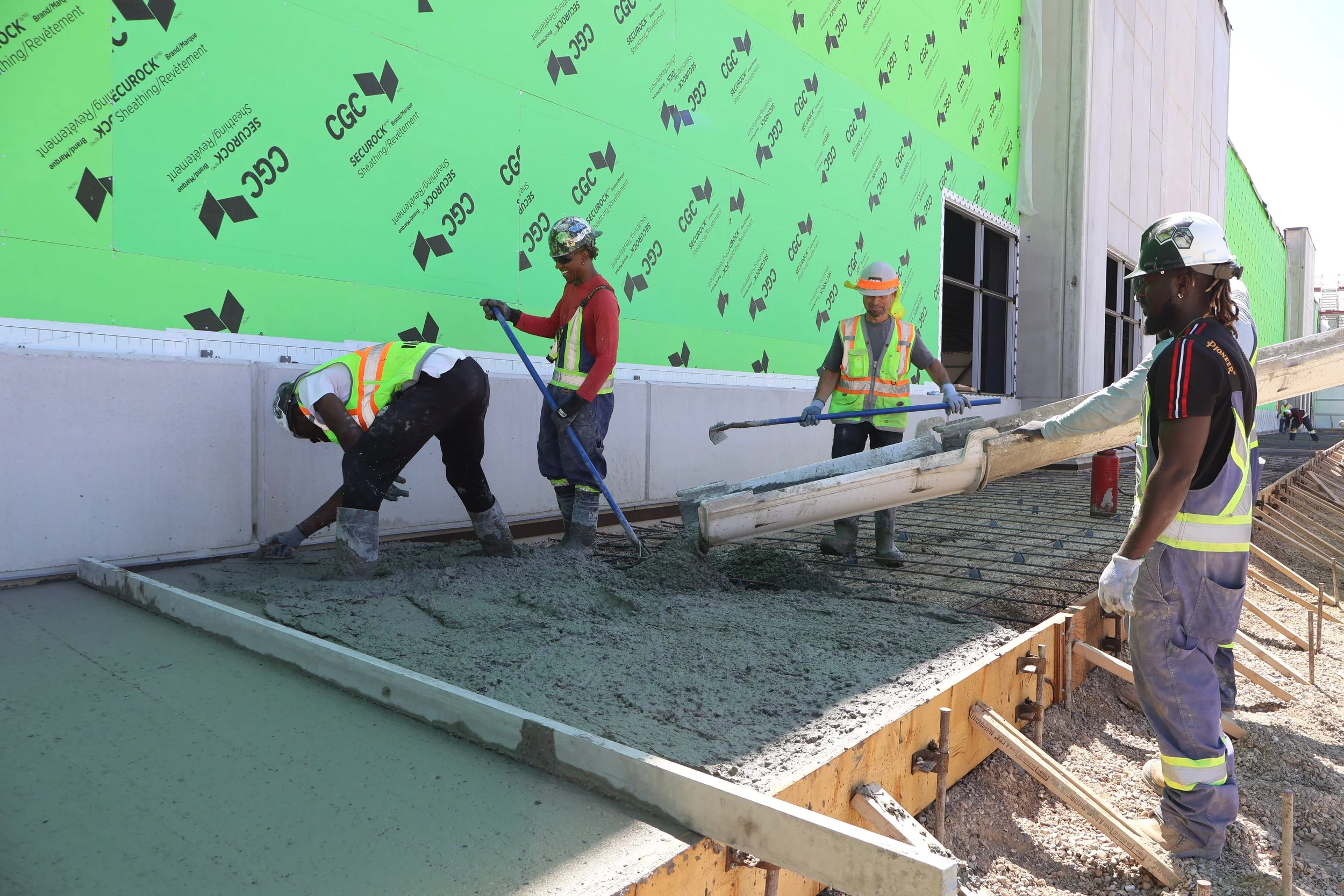 Construction workers wearing helmets and reflective vests working on pouring concrete on a building site with green sheathing on the exterior wall.