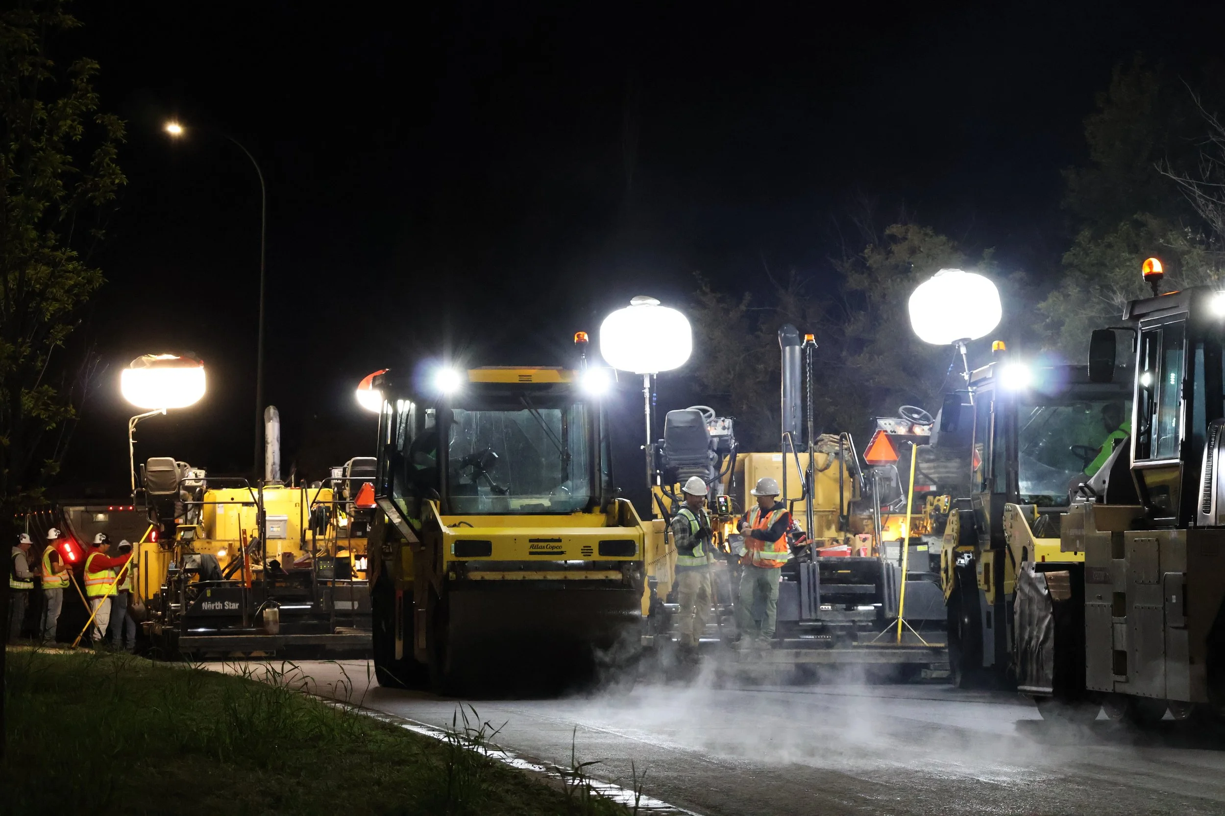 Construction crews working on a road at night with bright lights illuminating the scene, heavy machinery, and trees in the background.