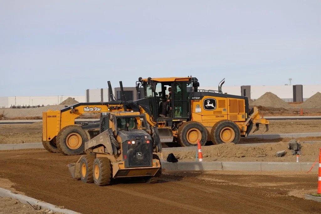 Construction site with two yellow John Deere construction vehicles working on dirt, with orange construction cones and a white noise barrier in the background.