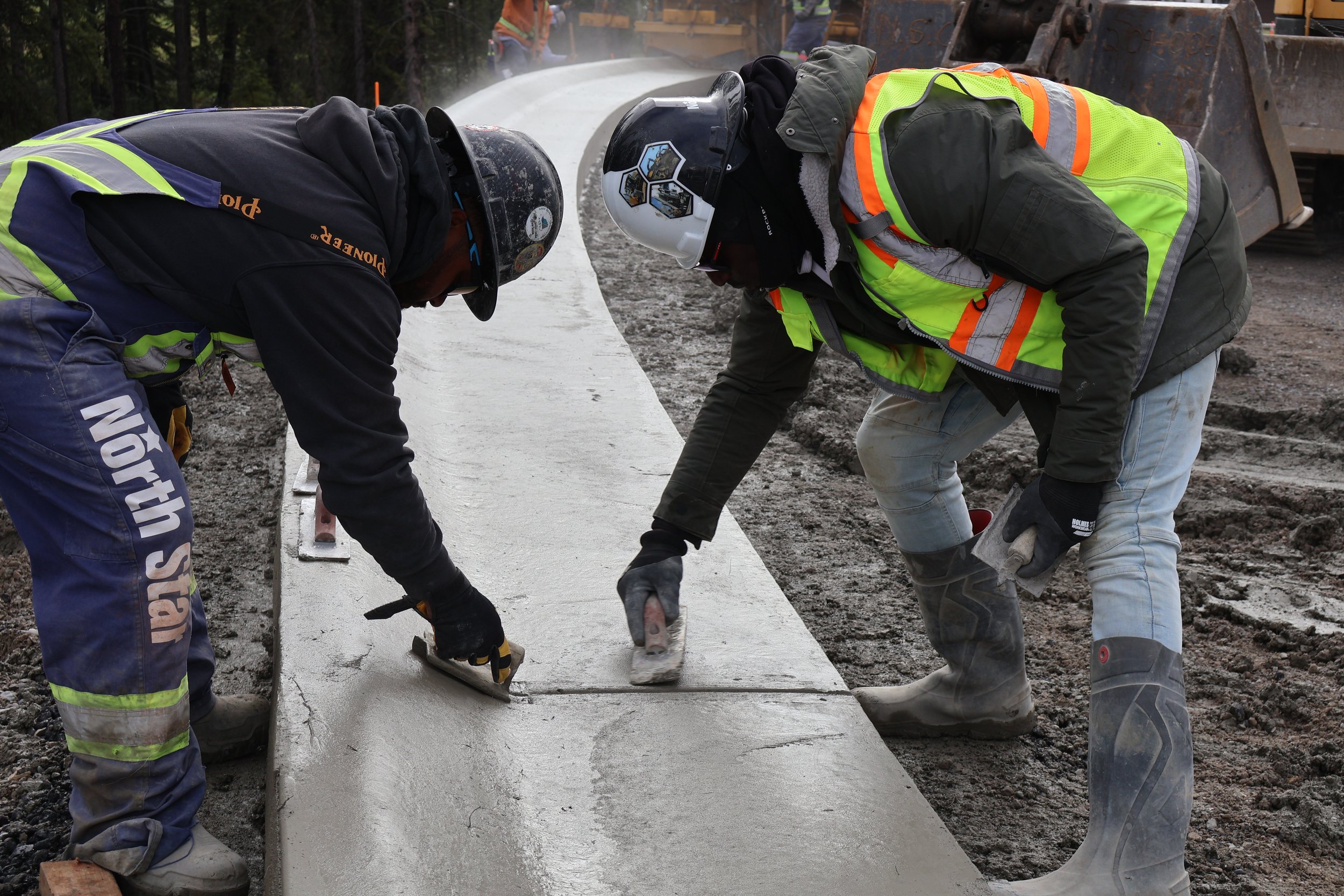 Two construction workers wearing safety helmets and reflective vests smoothing wet concrete on a road with construction machinery in the background.