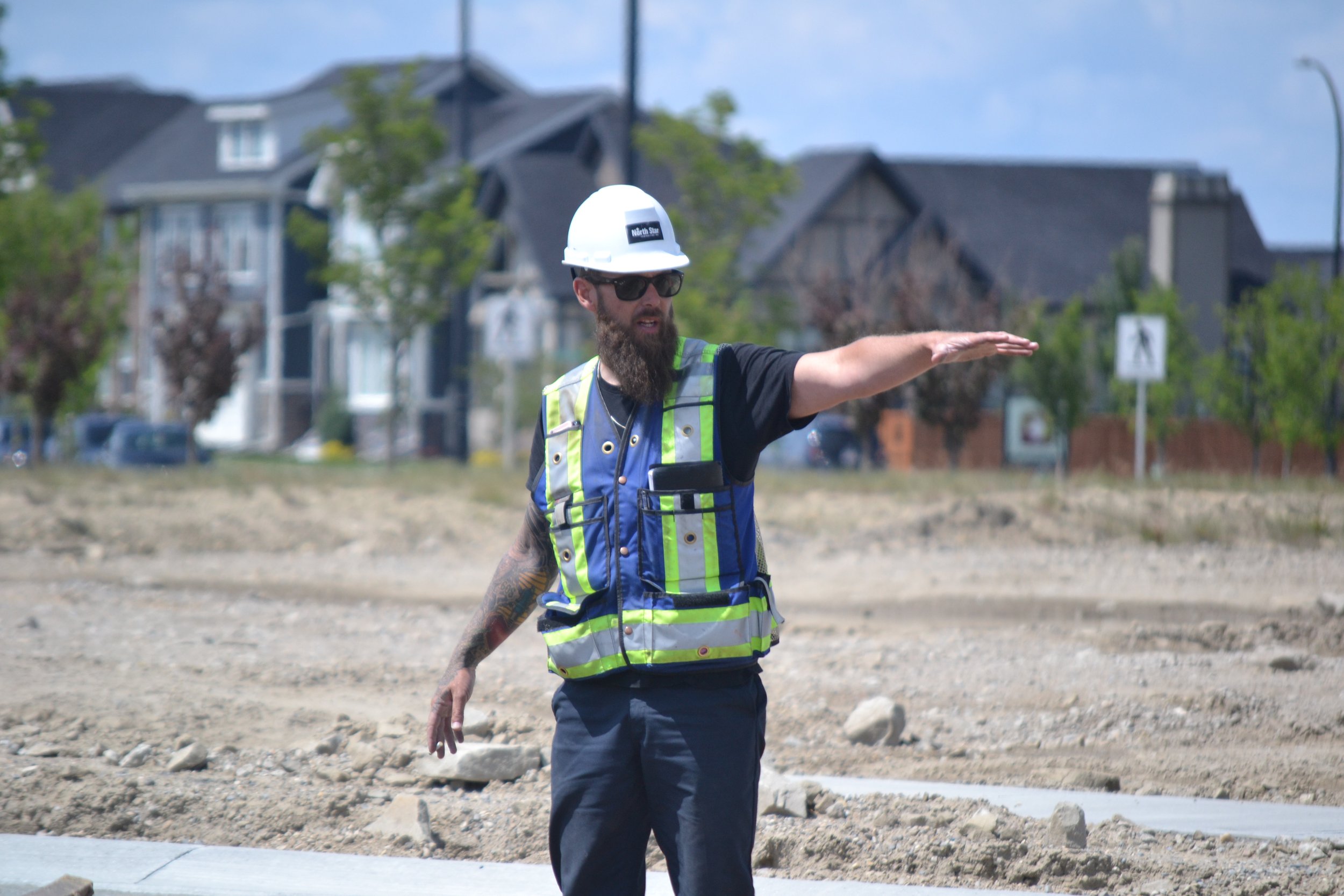 Construction worker with a beard and diamond-patterned tattoo on his arm, wearing sunglasses, a white hard hat labeled 'North Side,' a reflective safety vest, and dark clothing, gesturing with his right arm outstretched at an outdoor construction site with houses in the background.