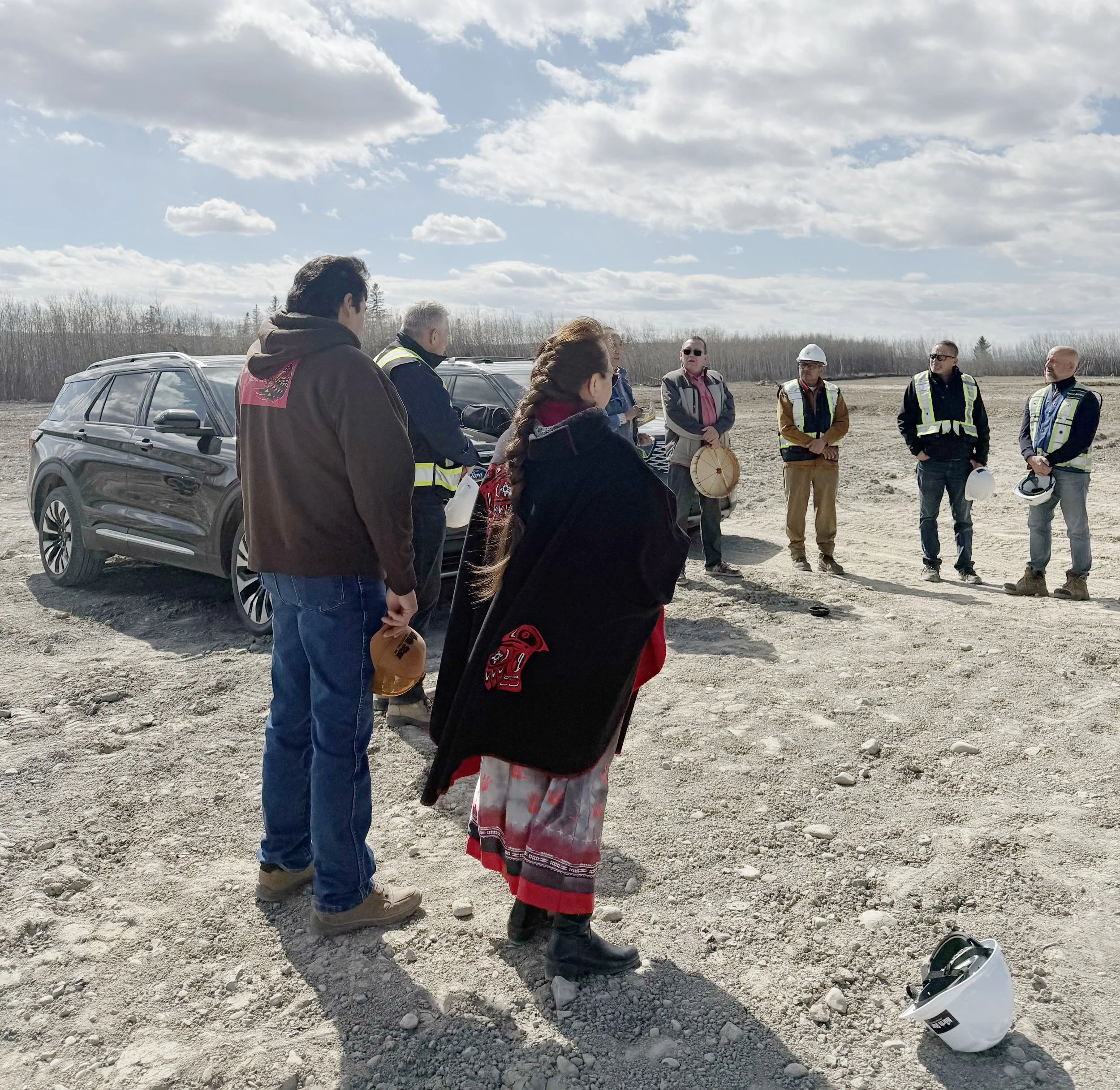 Group of people gathered outdoors on a cloudy day, some wearing construction vests and a hard hat, standing on a dirt surface near parked cars with a background of leafless trees and open sky.