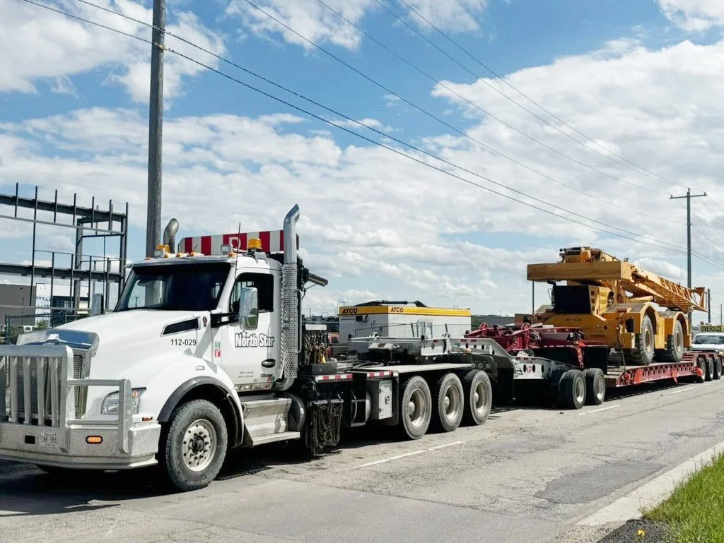 A large white truck carrying an oversized yellow crane on a red flatbed trailer parked on the side of a street.