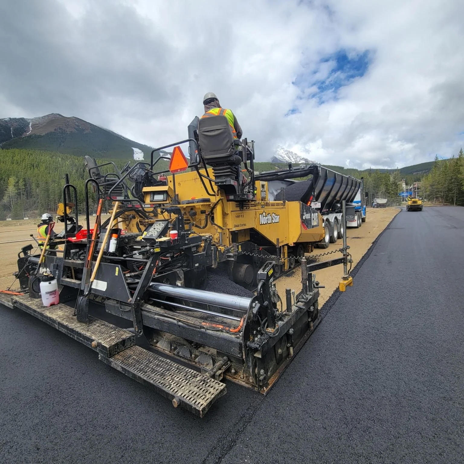 Construction workers operate a paving machine laying asphalt on a road with mountainous landscape in the background.