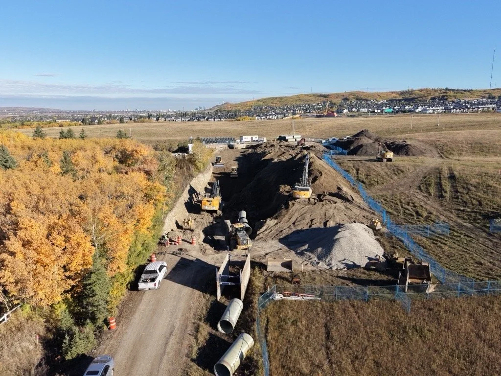 Construction site with excavators and trucks working on underground utilities or roadwork, surrounded by trees with fall foliage and open fields.