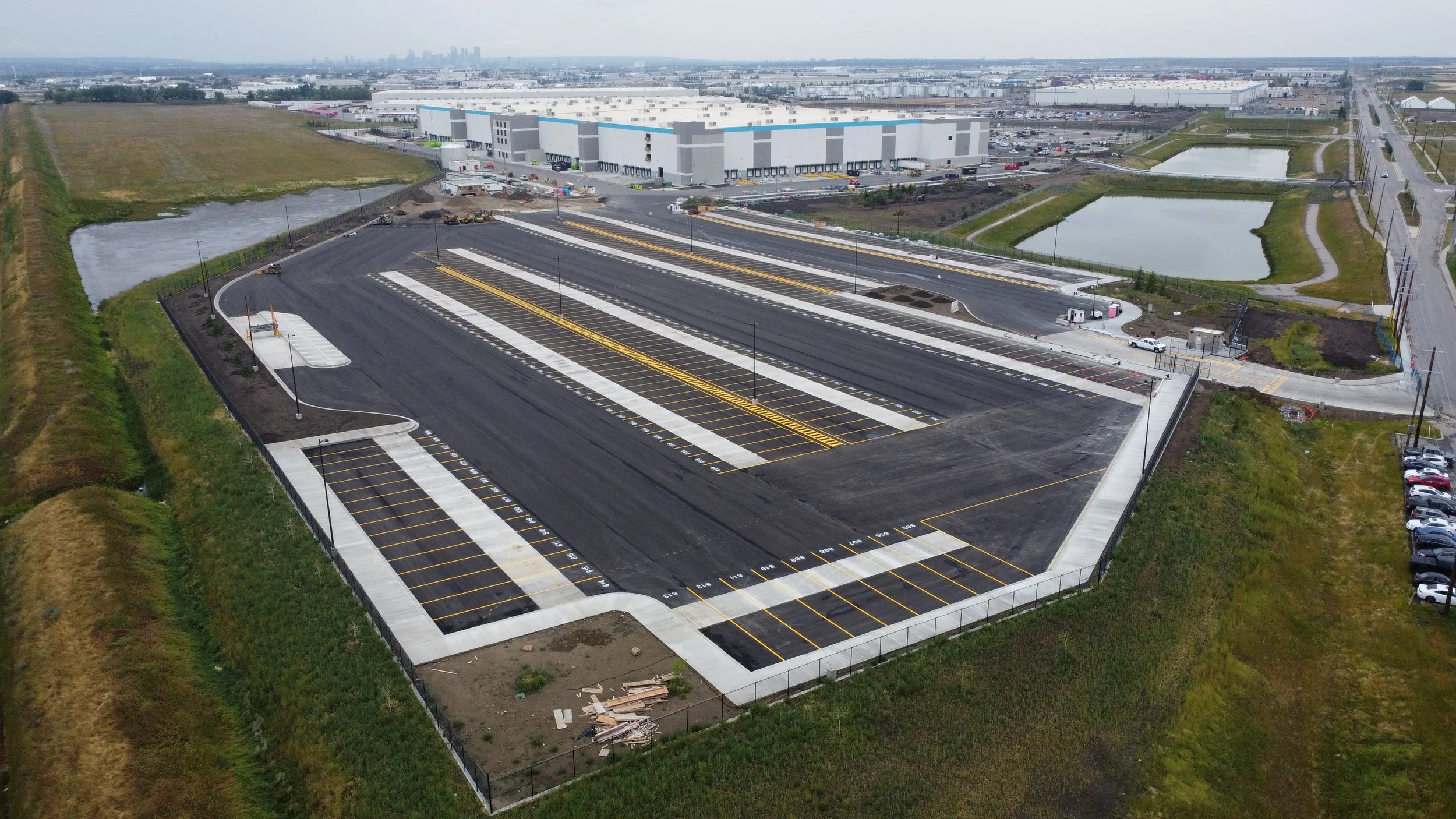 An aerial view of a large new parking lot with painted spaces, surrounded by grassy areas, near an industrial warehouse with loading docks, and some small ponds, with an urban skyline visible in the distance.