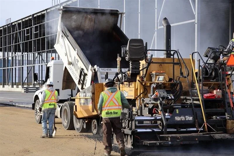 Construction workers in safety vests and helmets operate heavy machinery to lay asphalt on a road.