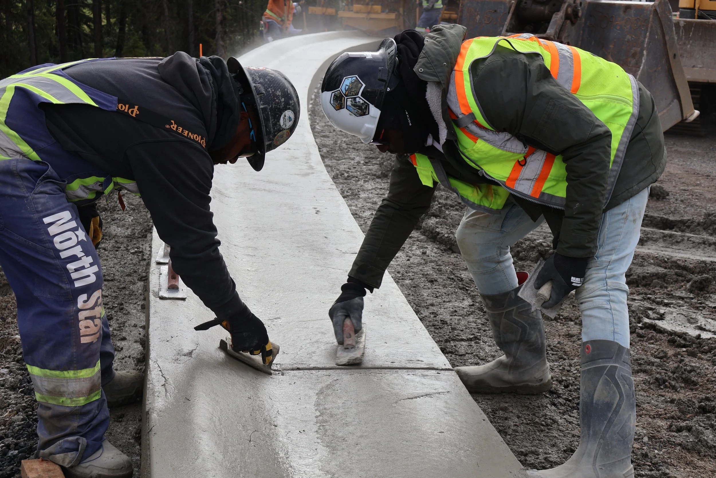 Two construction workers kneel on the ground, smoothing wet concrete on a sidewalk, with construction vehicles and workers in the background.