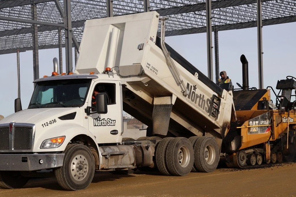 A large white North Star dump truck unloading asphalt mix into an industrial paving machine at a construction site.