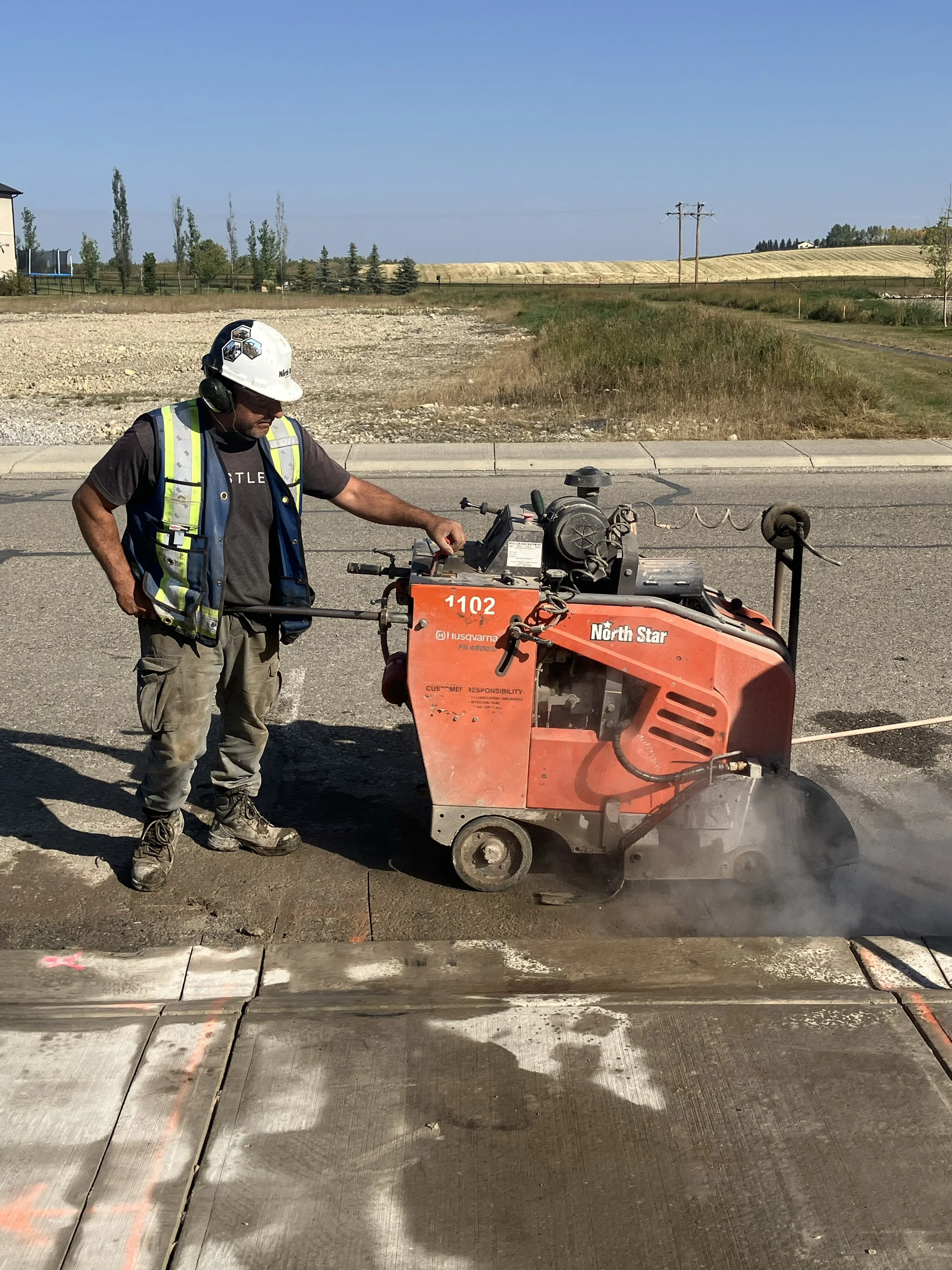 Worker wearing a white safety helmet, ear protection, and a reflective vest uses a concrete cutting machine on a street with freshly cut concrete slabs.