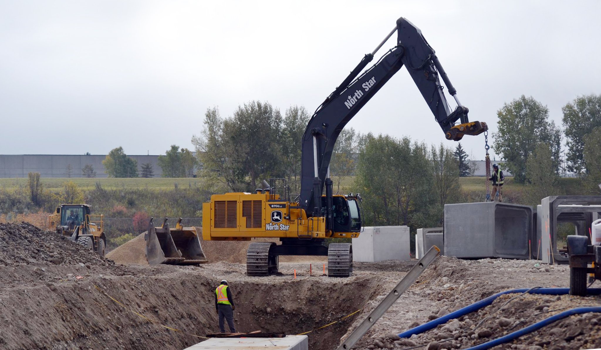 Construction site with bulldozer and excavator, workers, and concrete blocks, with trees and cloudy sky in the background.