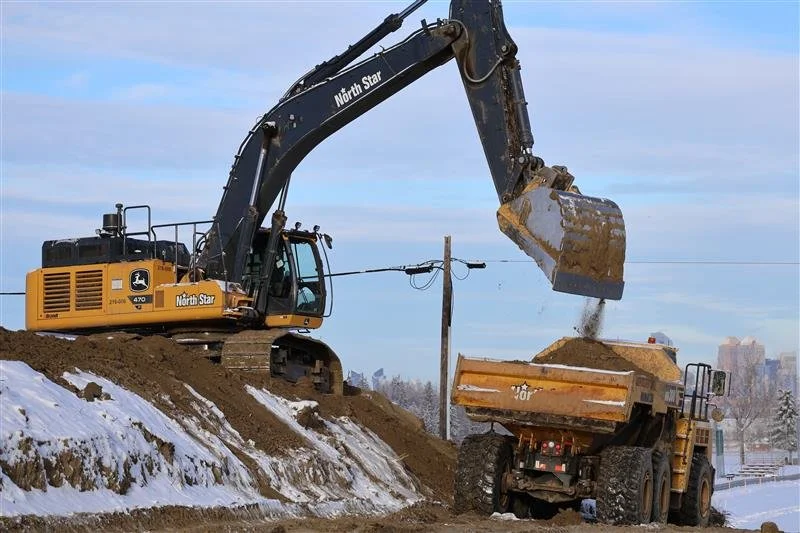 A John Deere excavator loading dirt into a yellow dump truck at a construction site with snow on the ground and city buildings in the distance.