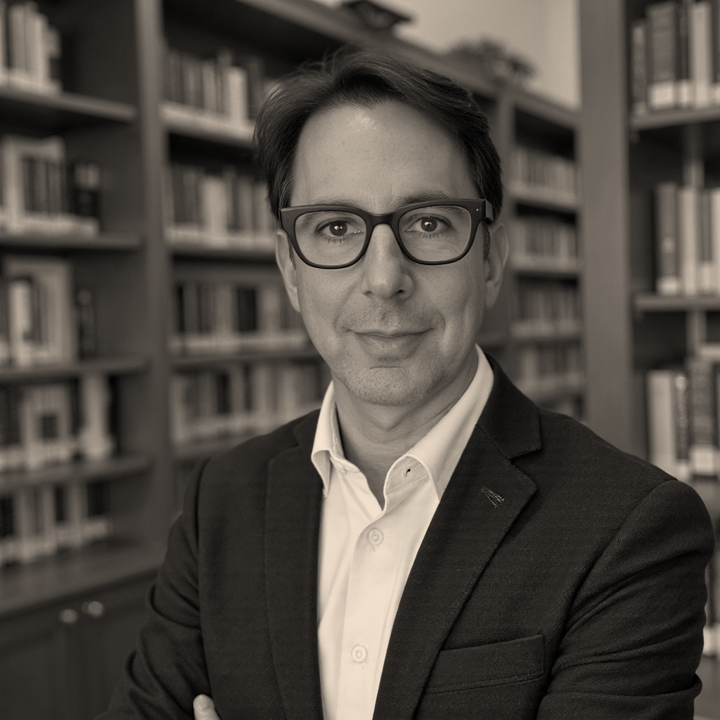 A man in a suit and glasses standing in front of bookshelves in a library.