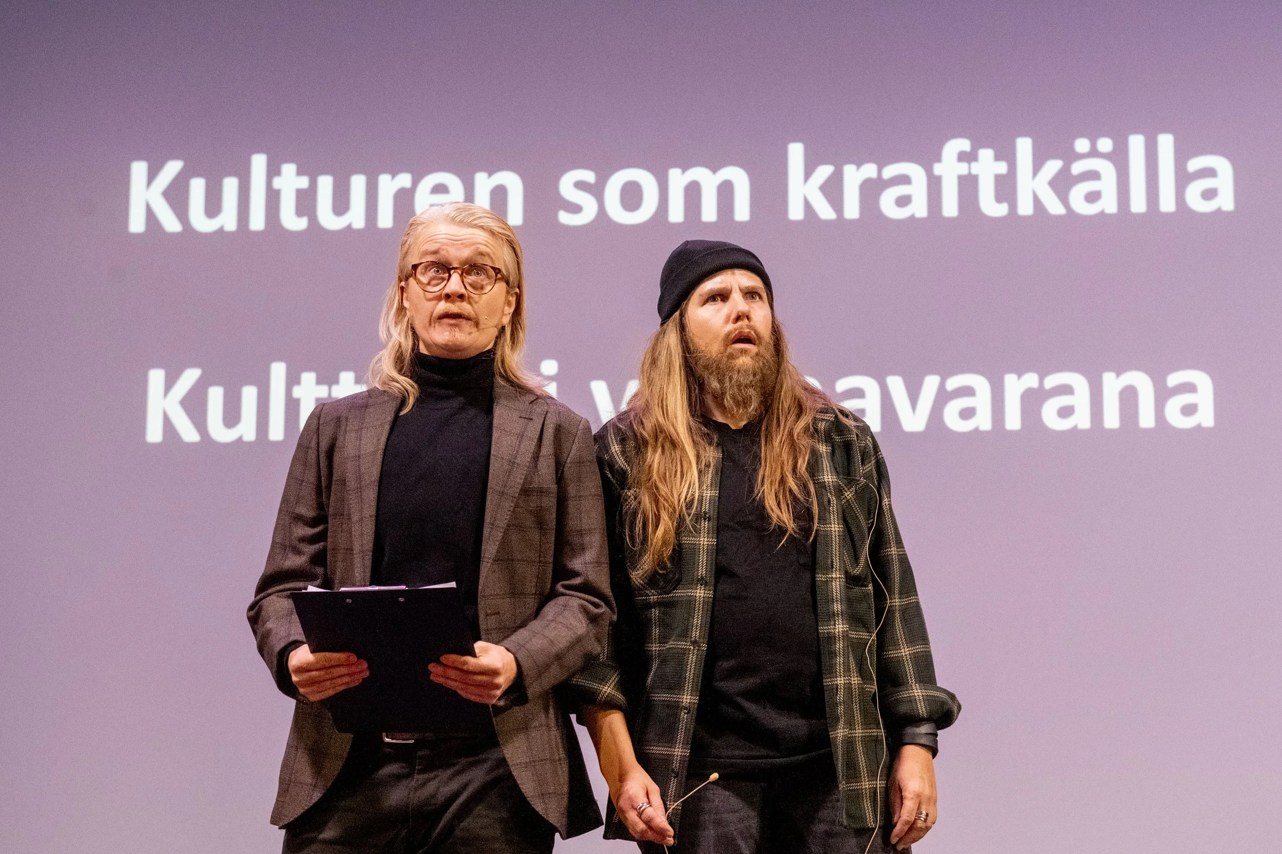 Two people standing in front of a screen with Swedish text, one holding a clipboard, both looking surprised or confused.