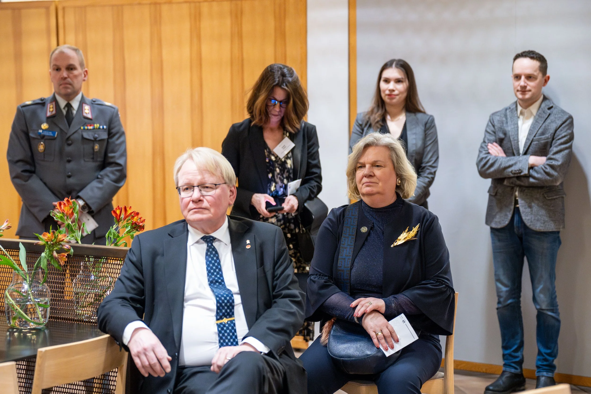 A group of five people in formal attire, two seated in the foreground and three standing behind them, in a wood-paneled room, possibly attending a formal event or meeting.