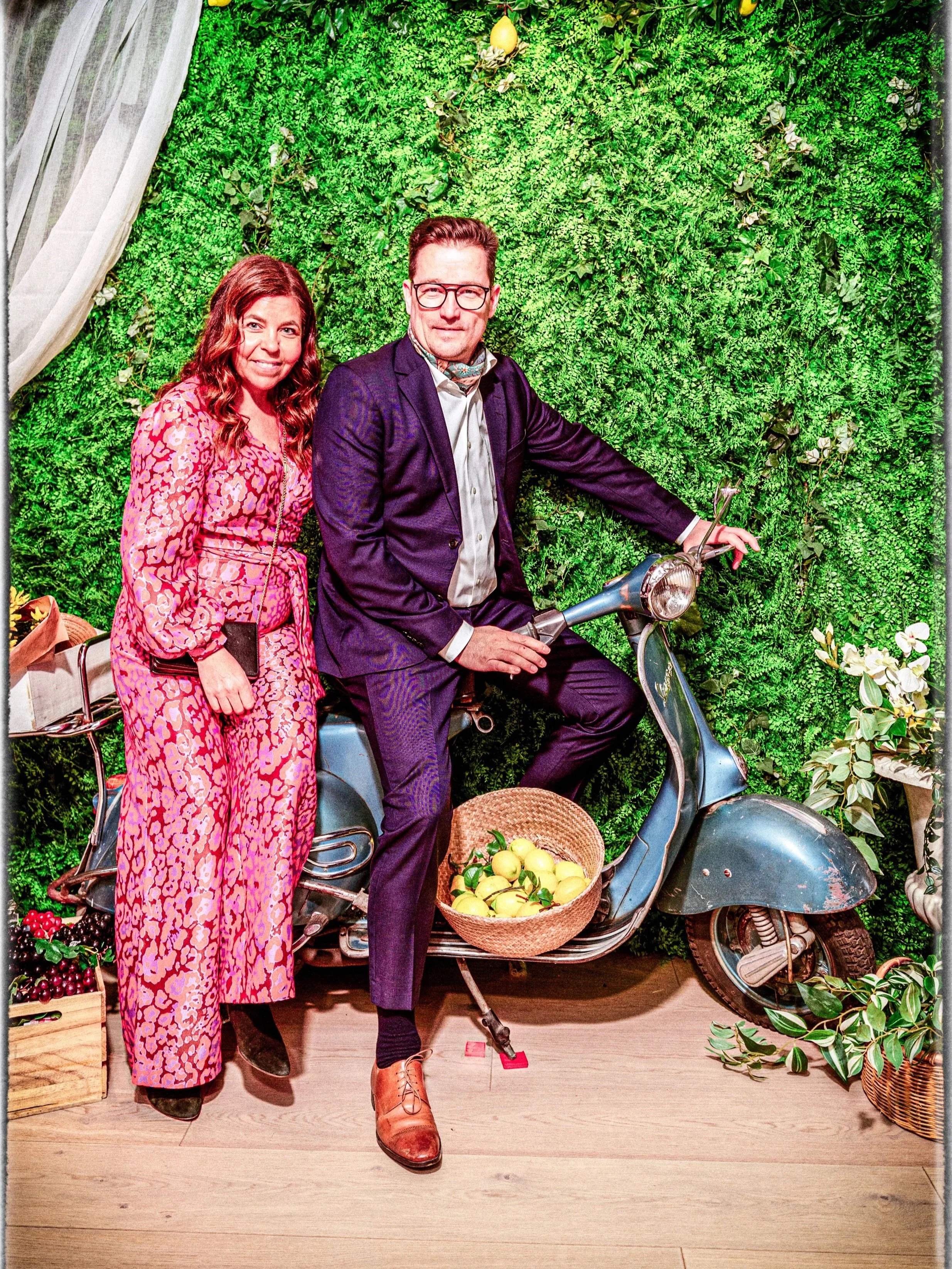 A man and woman posing next to a vintage scooter with a basket of yellow apples. The woman is wearing a pink patterned jumpsuit and holding a phone, while the man is sitting on the scooter wearing a dark suit and glasses, with a green leafy wall back