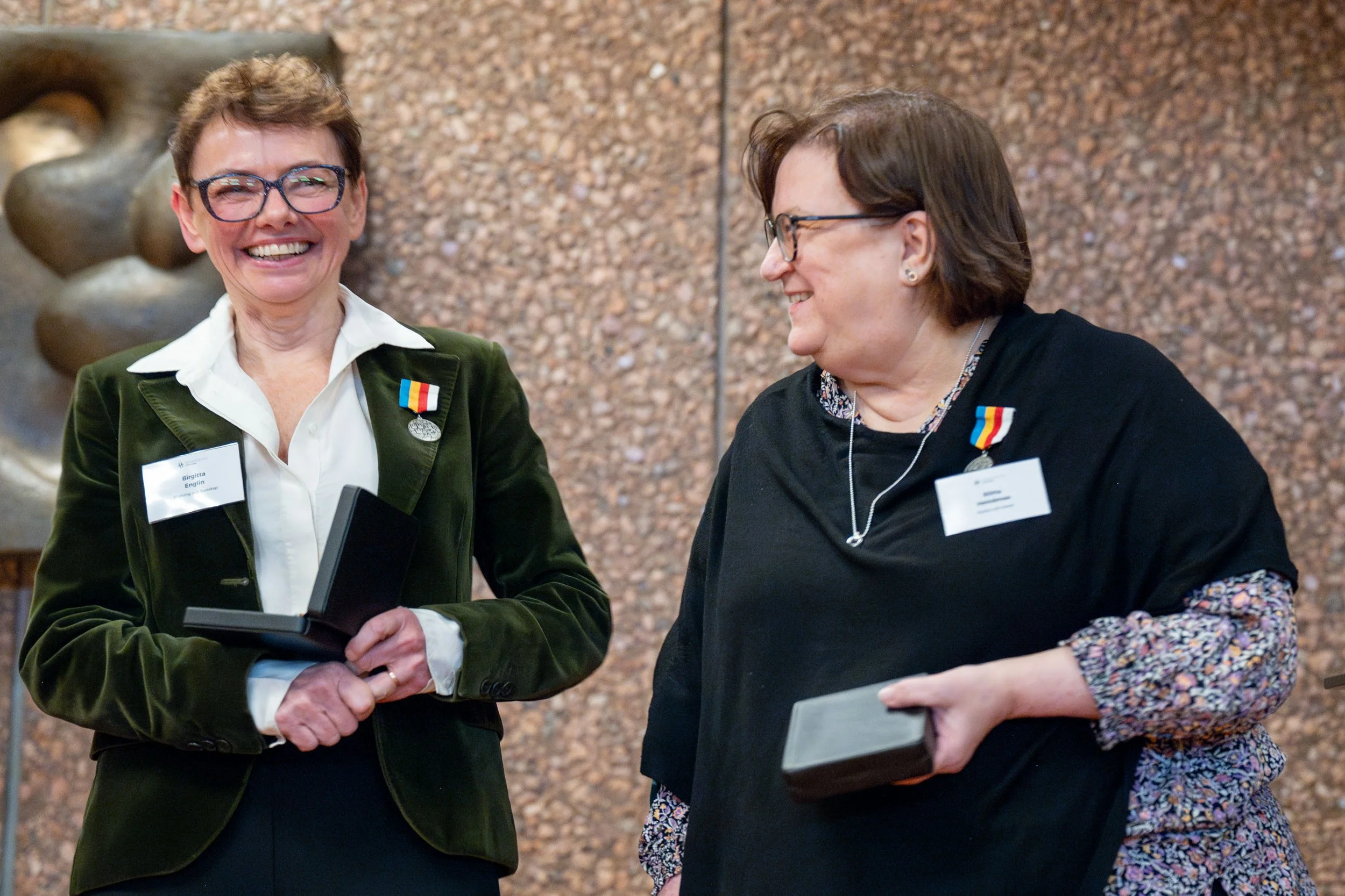 Two women wearing glasses are sharing a joyful moment, holding awards or folders, with medals on their clothing, standing in front of a textured wall.