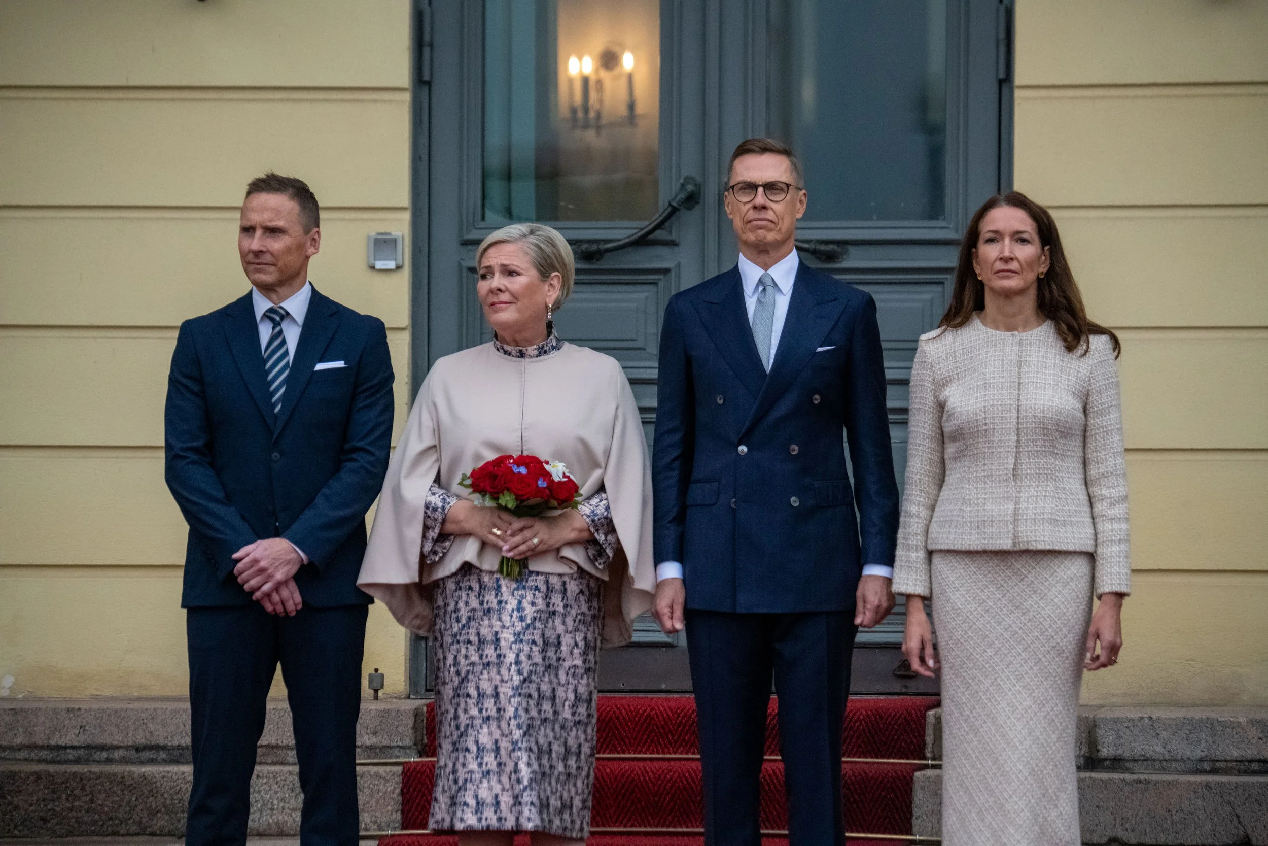 Four people standing on red carpet outside a building, dressed formally; one woman holding a bouquet of red flowers.