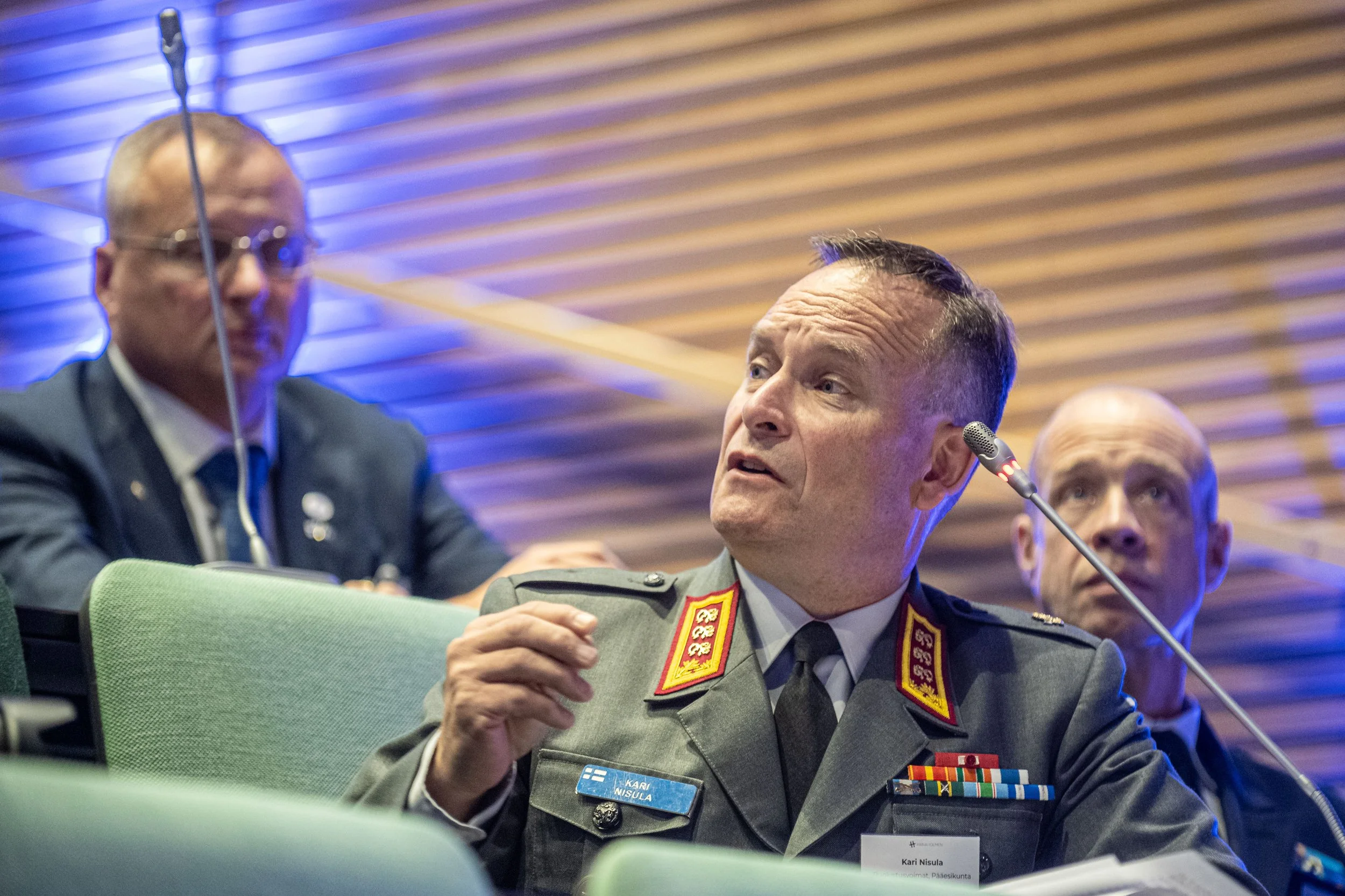 A man in a military uniform with medals and a name tag during a meeting or conference, engaging in conversation, with two other men in the background, one in a suit and the other in a uniform, all seated at a table with microphones.