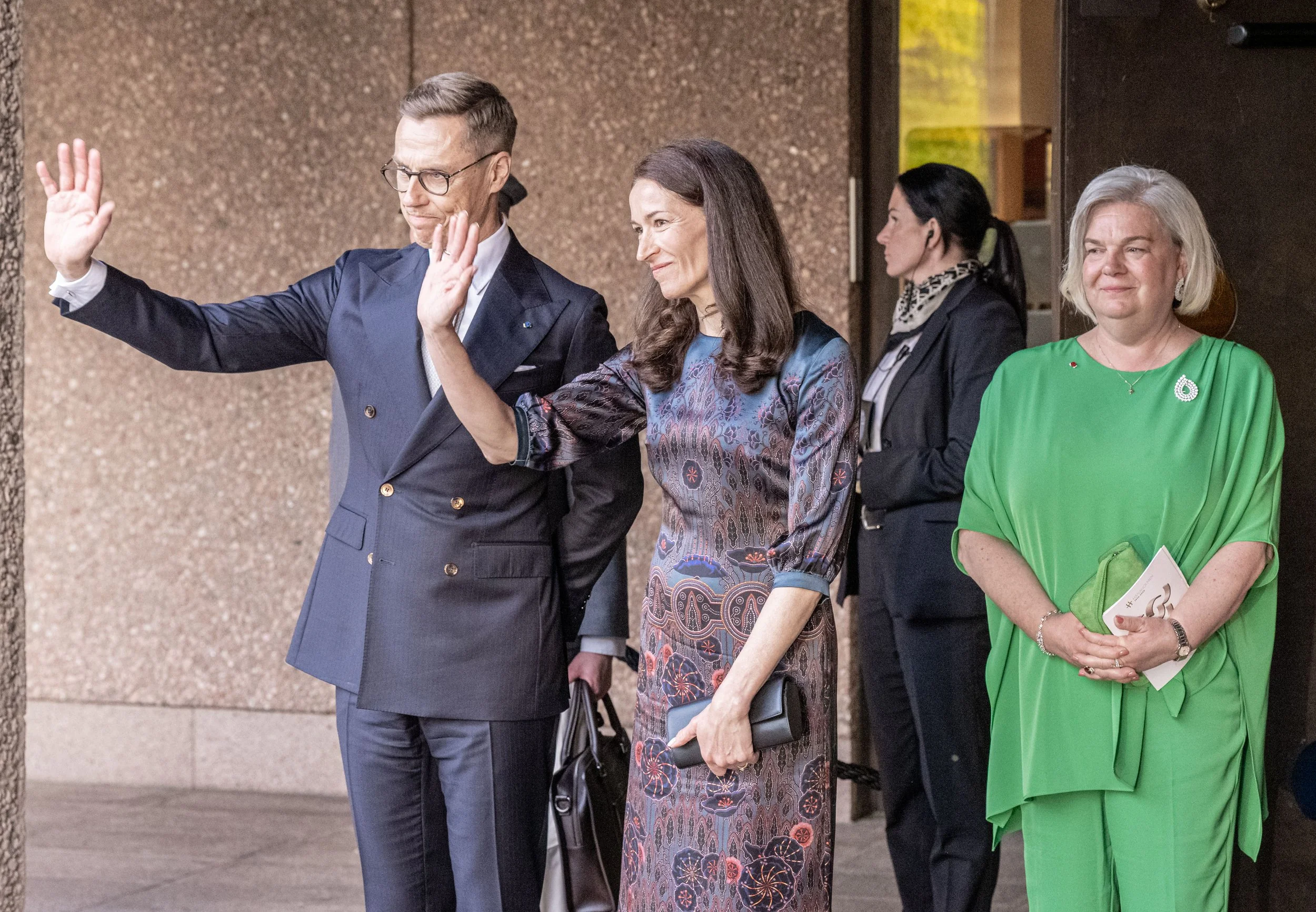 Four formally dressed women and one man with glasses, standing outside a building, waving and holding items, during a formal event.