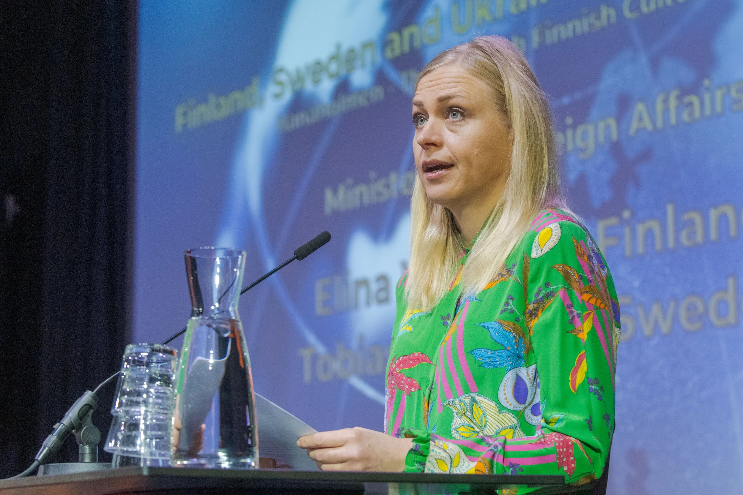A woman with blonde hair wearing a colorful, patterned green dress stands at a podium, speaking into a microphone, with a blue background featuring text related to Finland, Sweden, and other European countries.