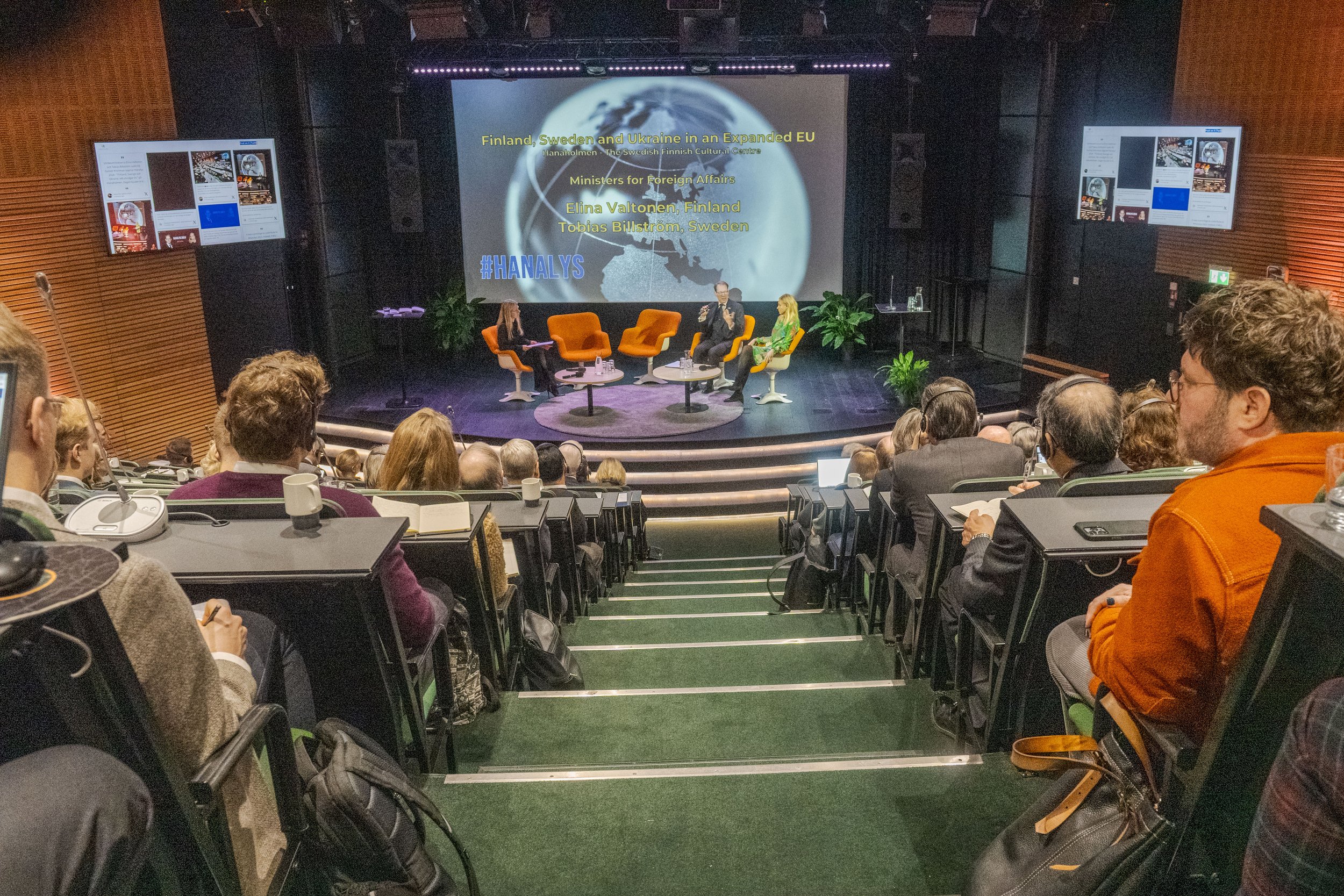 A panel discussion taking place in a conference hall with multiple attendees. Four people are seated on stage with a large screen behind them displaying text about Finland, Sweden, Ukraine, and the EU. The audience is seated in rows, some taking note