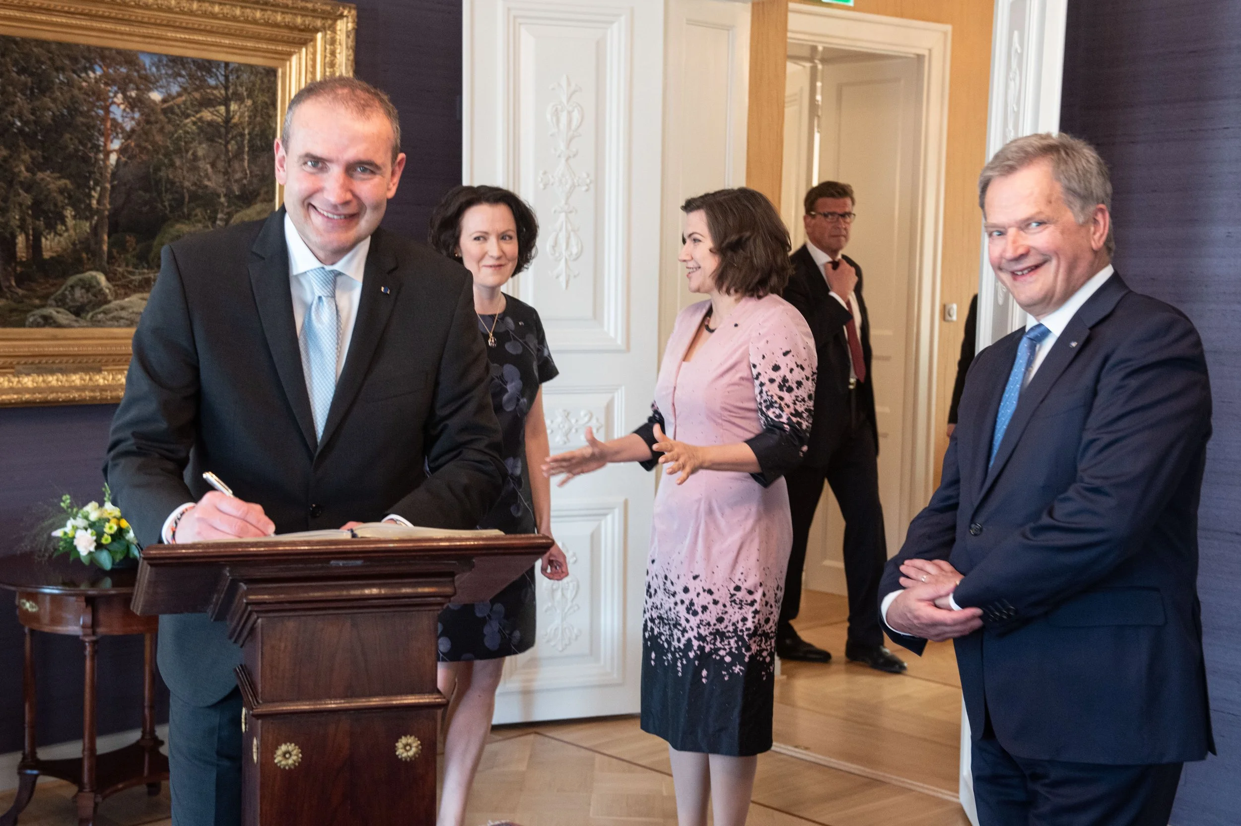 A man signing a document at a formal event with four other well-dressed individuals standing around him, in a room with elegant decor and a large framed painting.