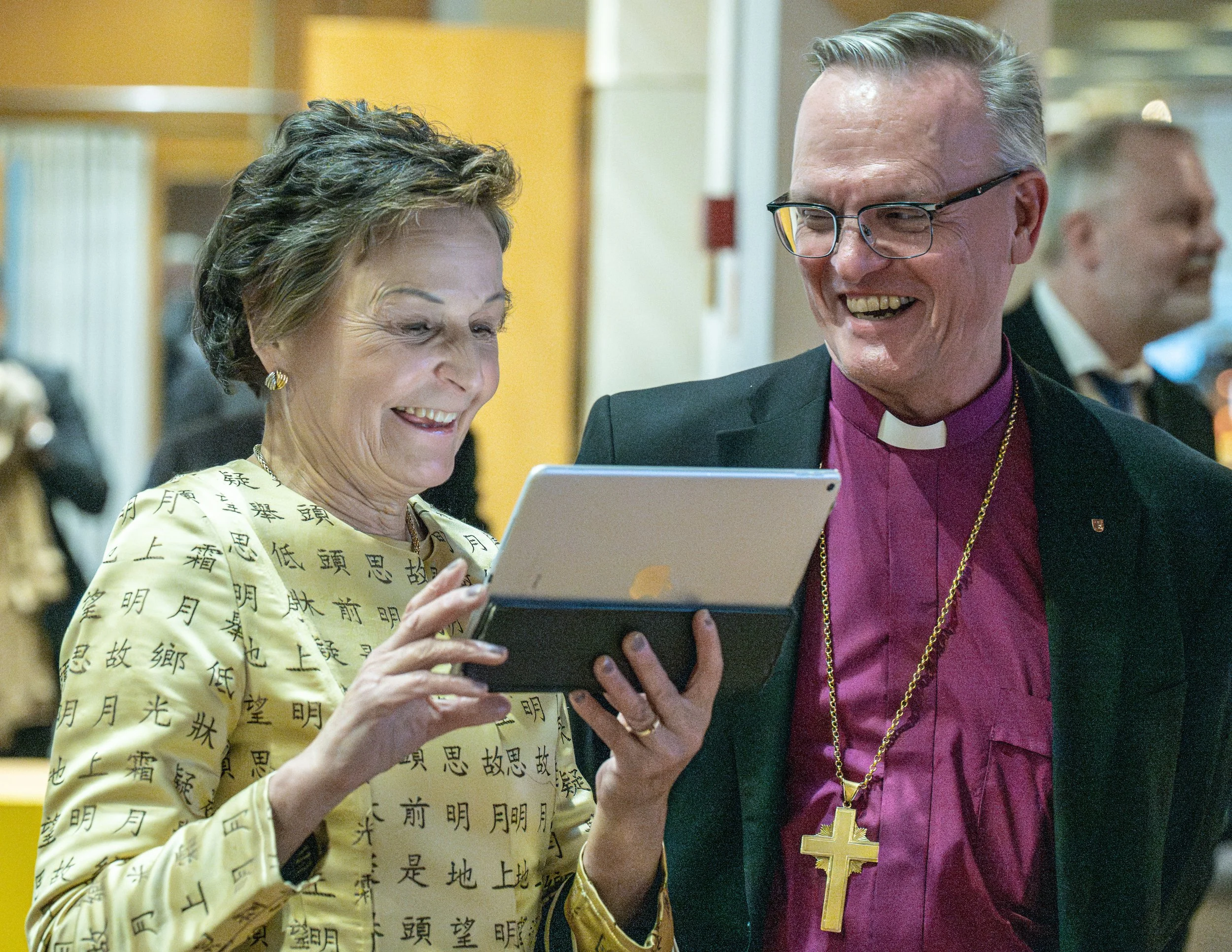 A woman and a man, likely a bishop, are smiling and looking at a tablet device together. The woman is wearing a light-colored outfit with Asian characters printed on it, and the man is dressed in church attire with a purple shirt, a black jacket, and